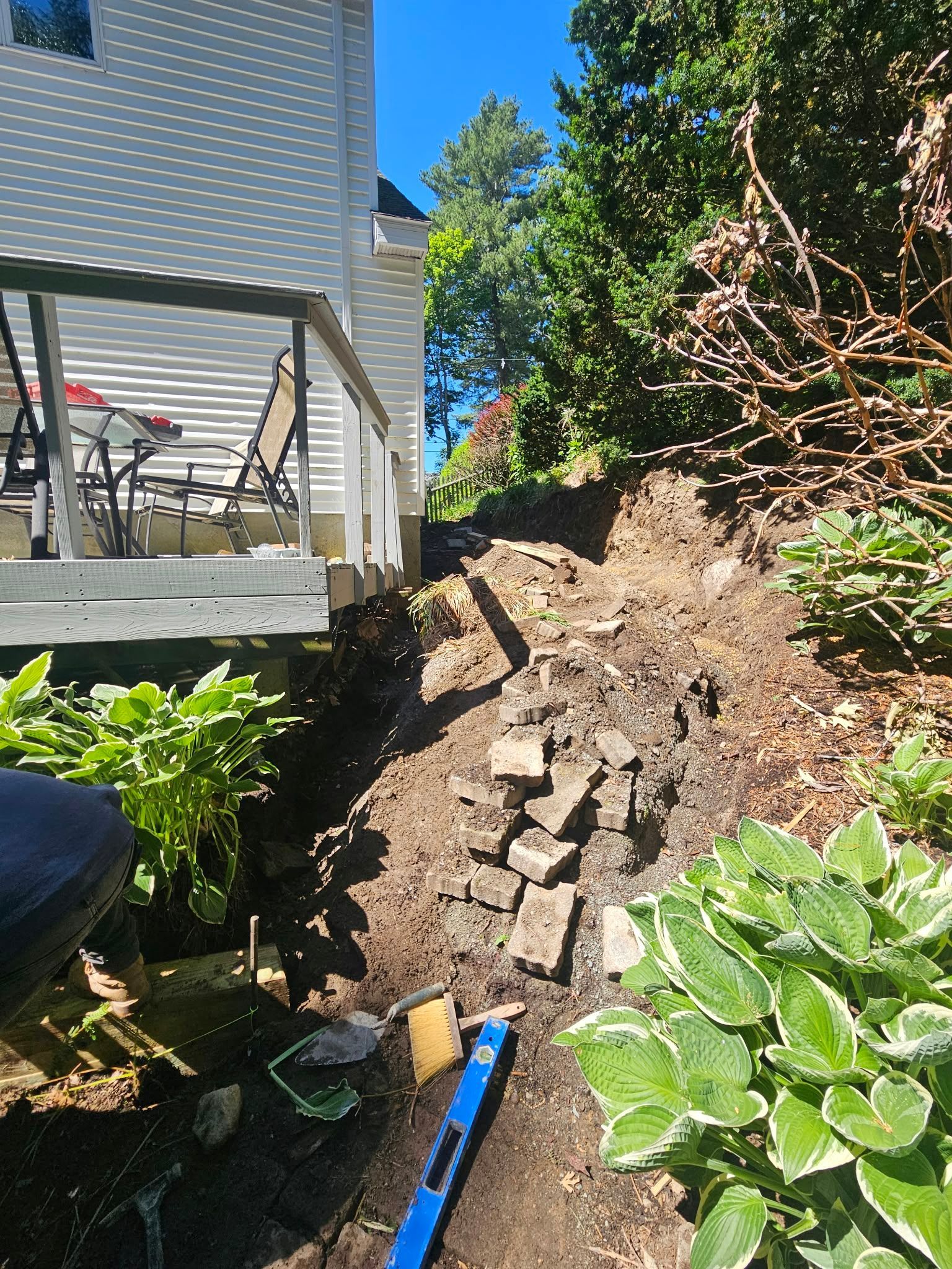 Backyard hillside with exposed soil and stacked pavers beside a porch and green plants