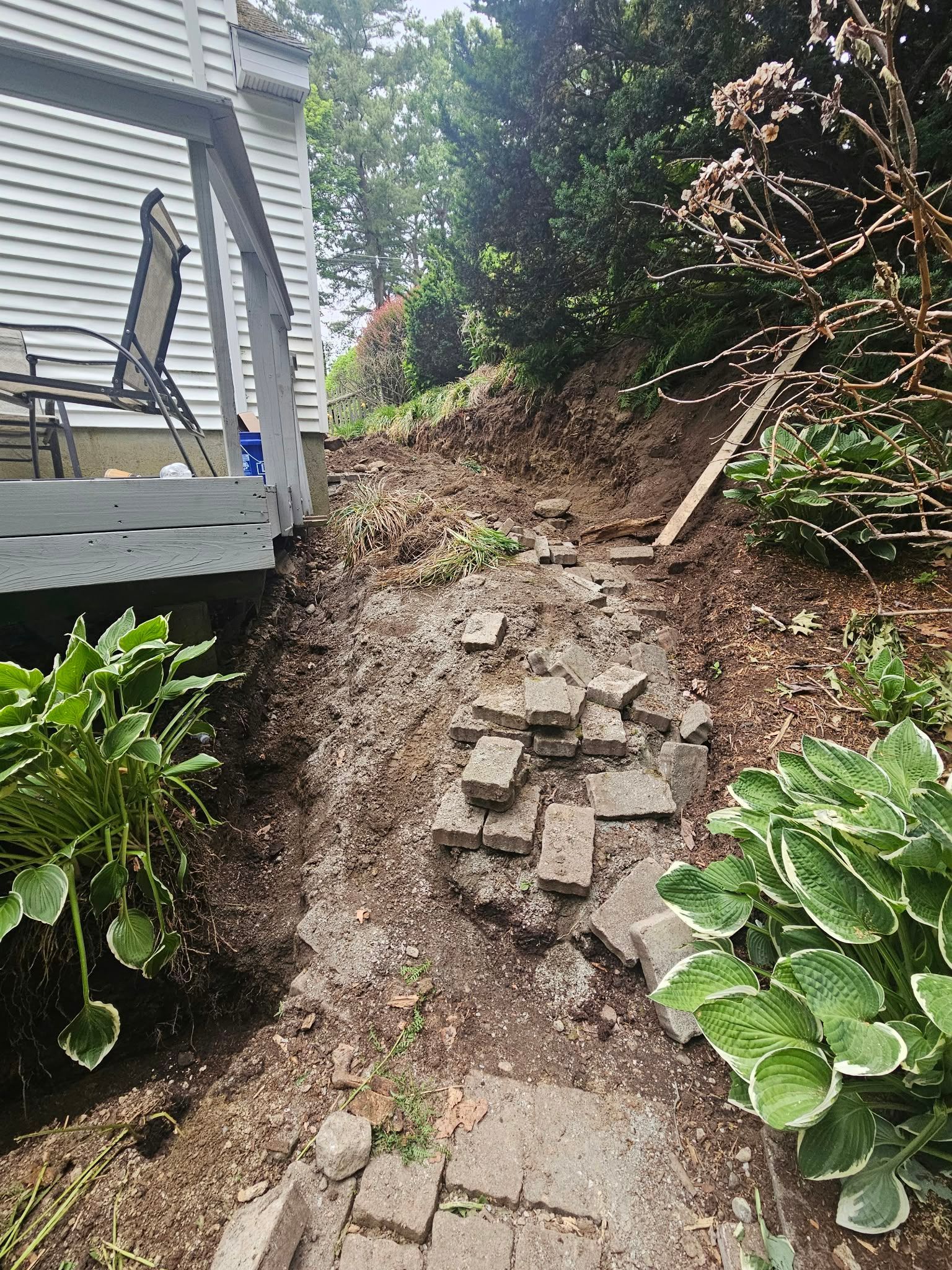 Steep dirt slope with loose rocks beside a house, lined by plants and a rough stone path.