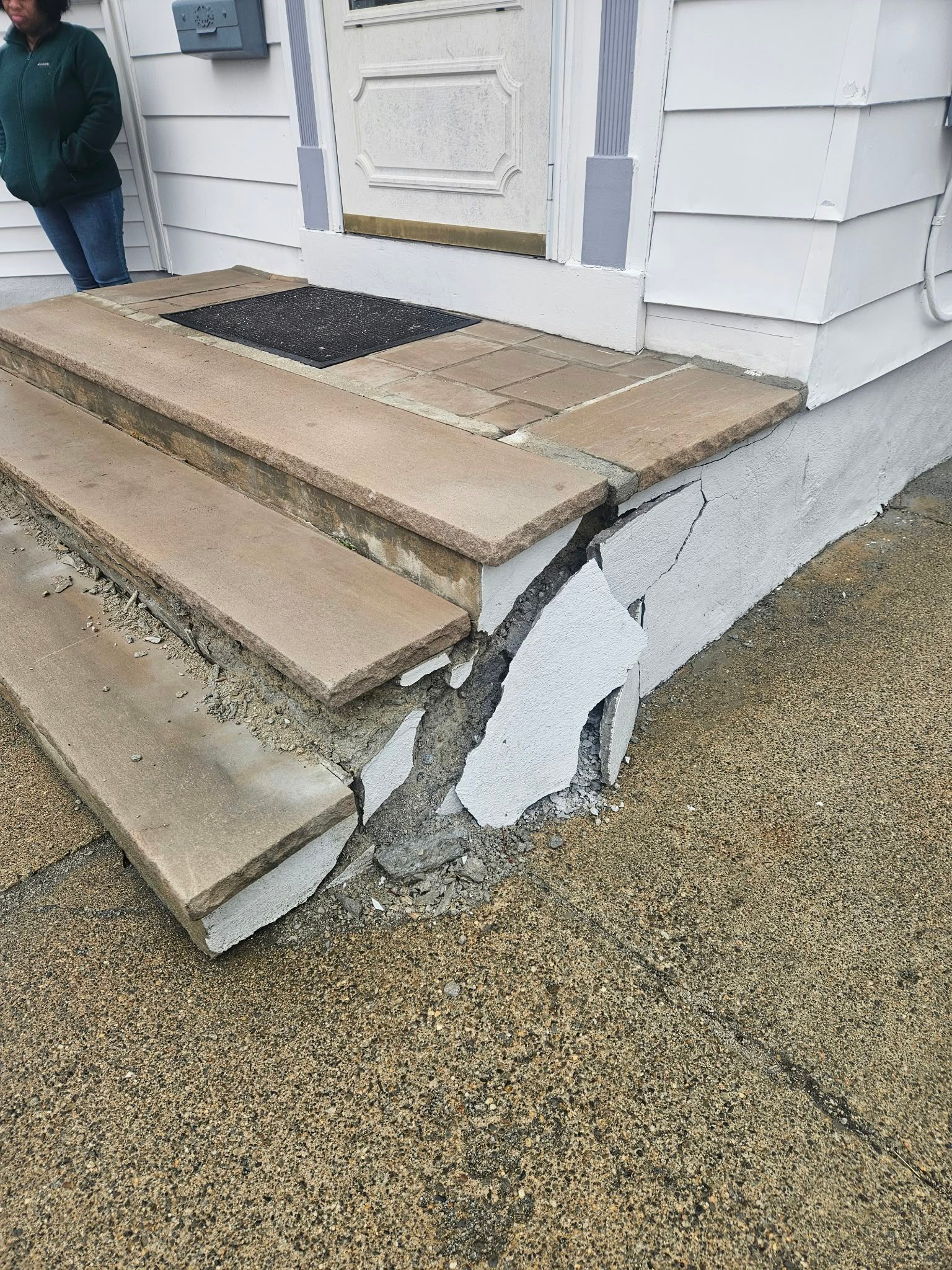 Damaged front porch steps with cracked concrete and chipped white siding beside a house entrance