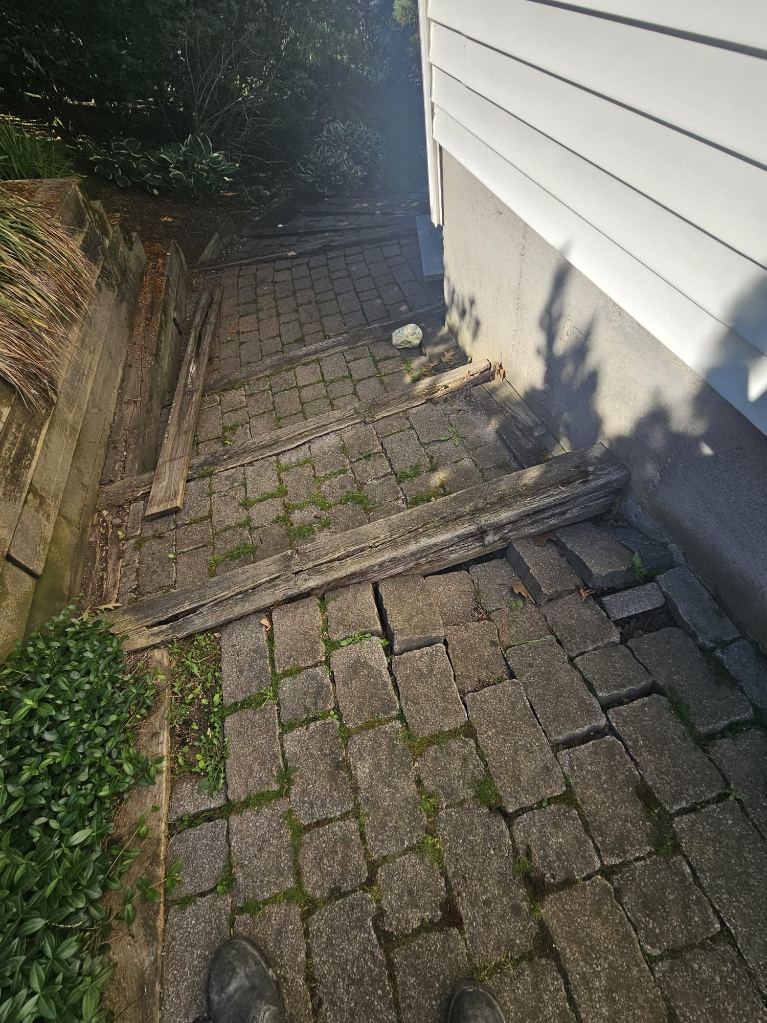 Narrow brick-paved side yard beside a white house, with retaining wall and greenery.