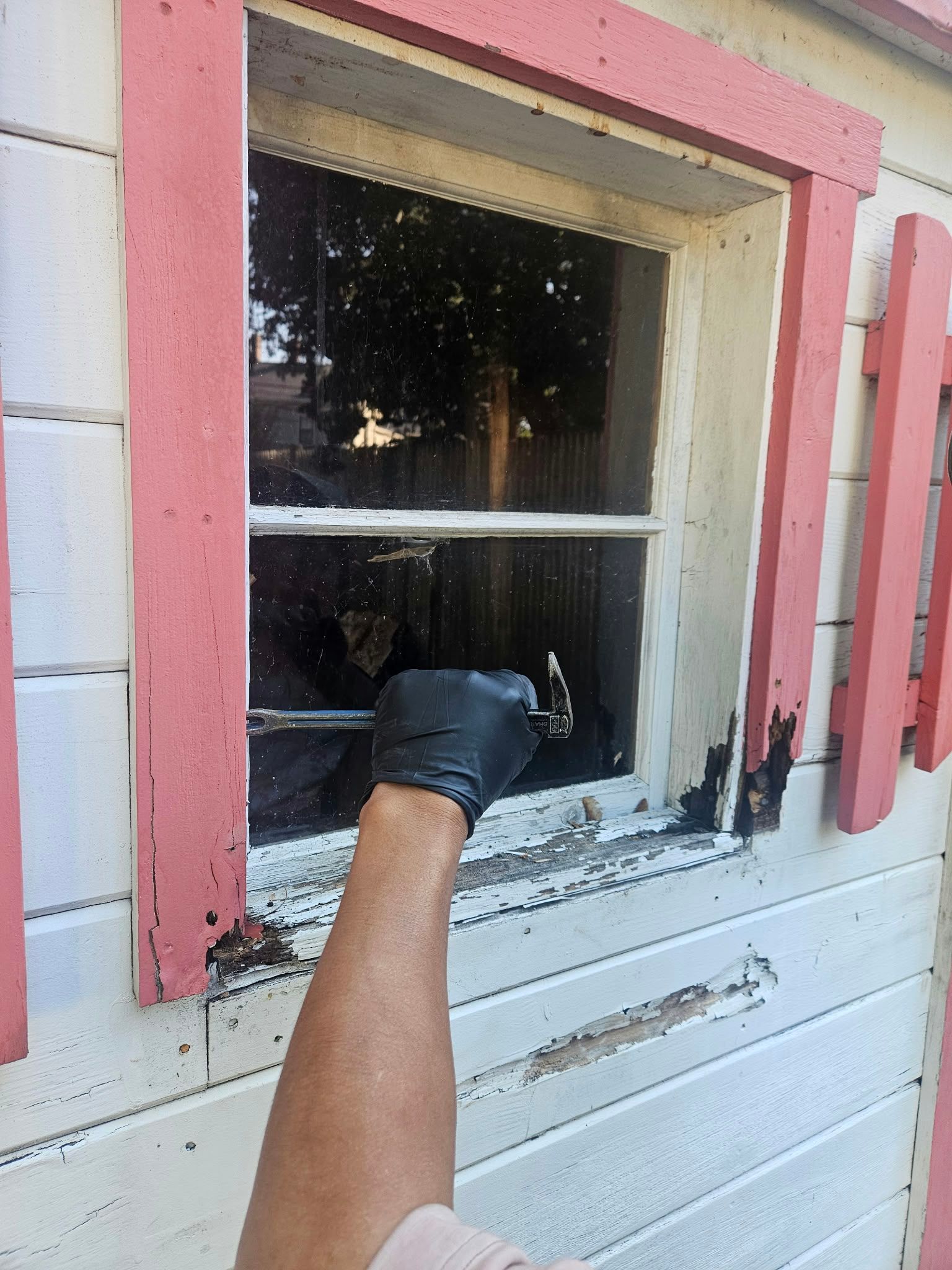 Gloved hand scraping white paint from a pink-trimmed window on a house exterior