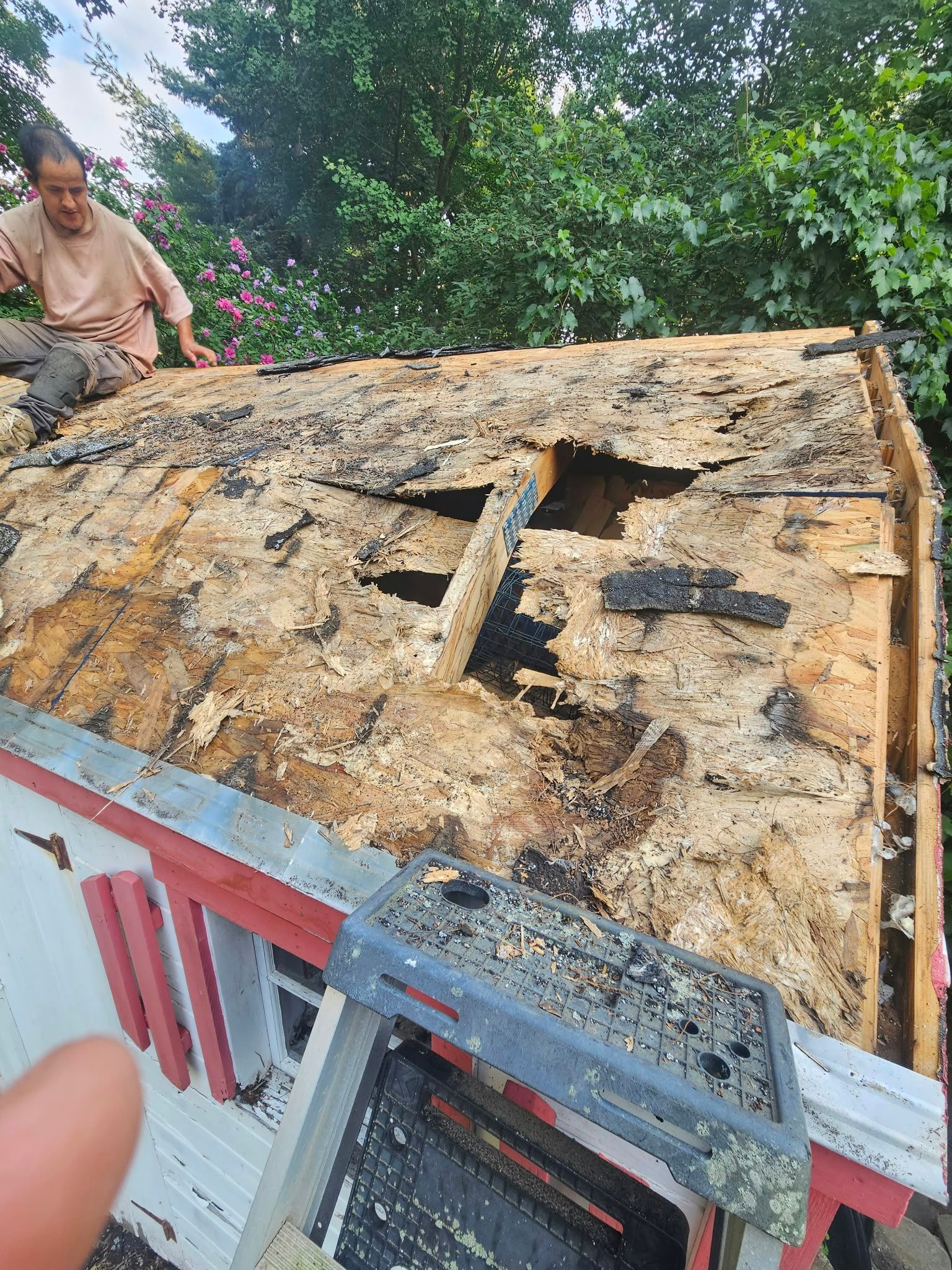 Damaged roof with exposed plywood and black roofing material; a person stands beside it outdoors.