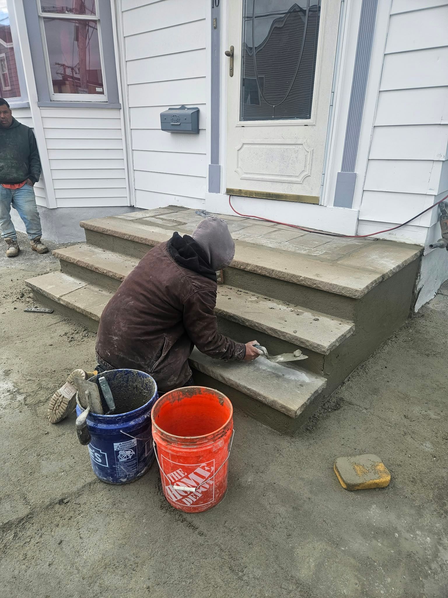 Worker painting concrete steps beside a white house, with blue and orange paint buckets.