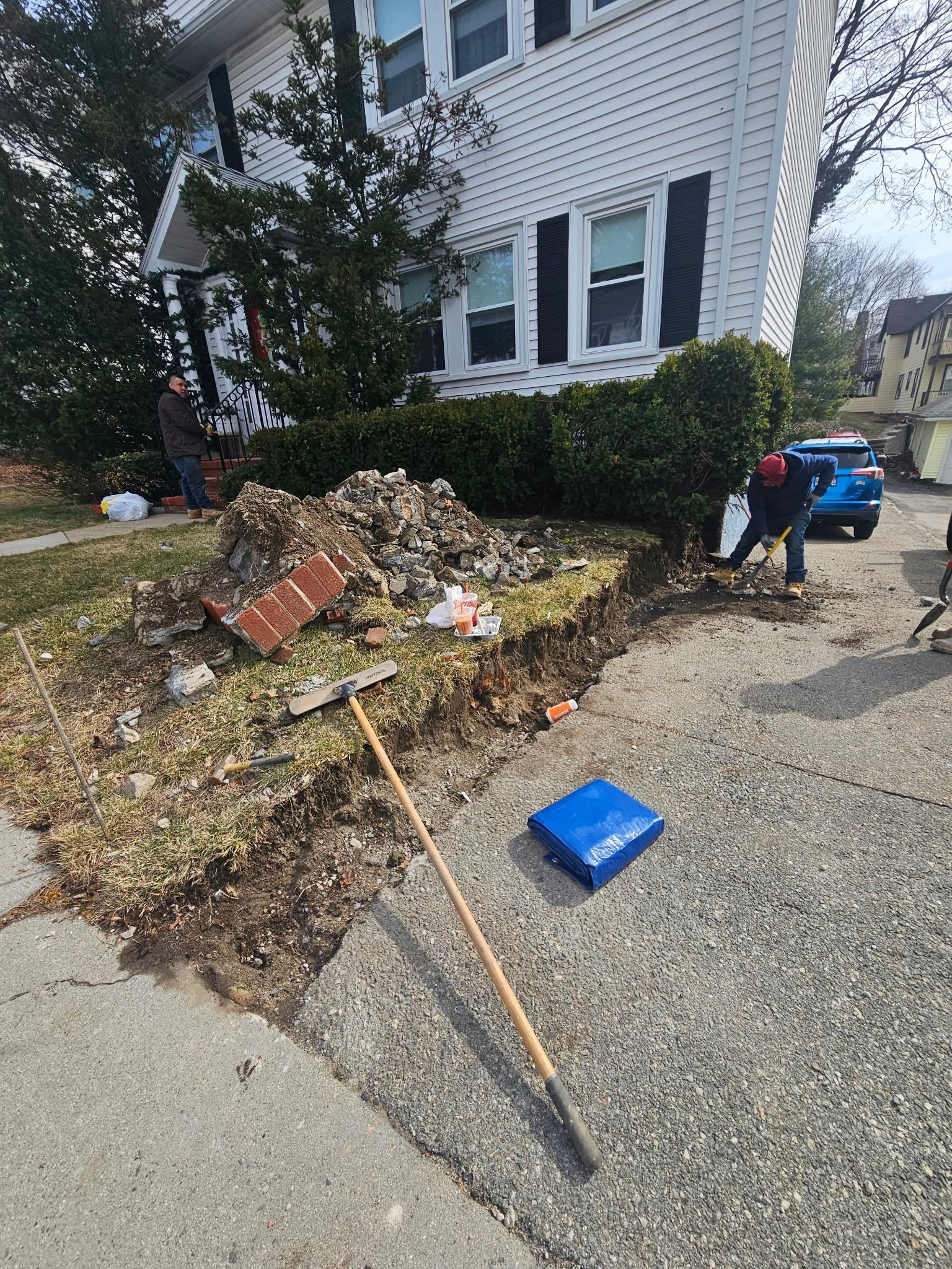 Large pile of yard waste beside a white house, with a rake and blue bin on the sidewalk.