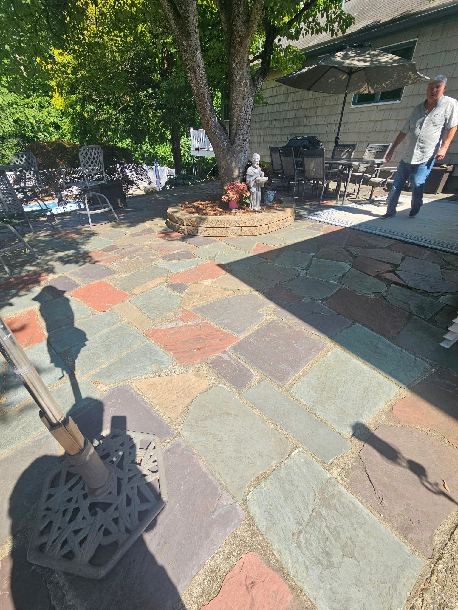 Patio with multicolored stone pavers, patio furniture, and a person standing by a tree in sunlight