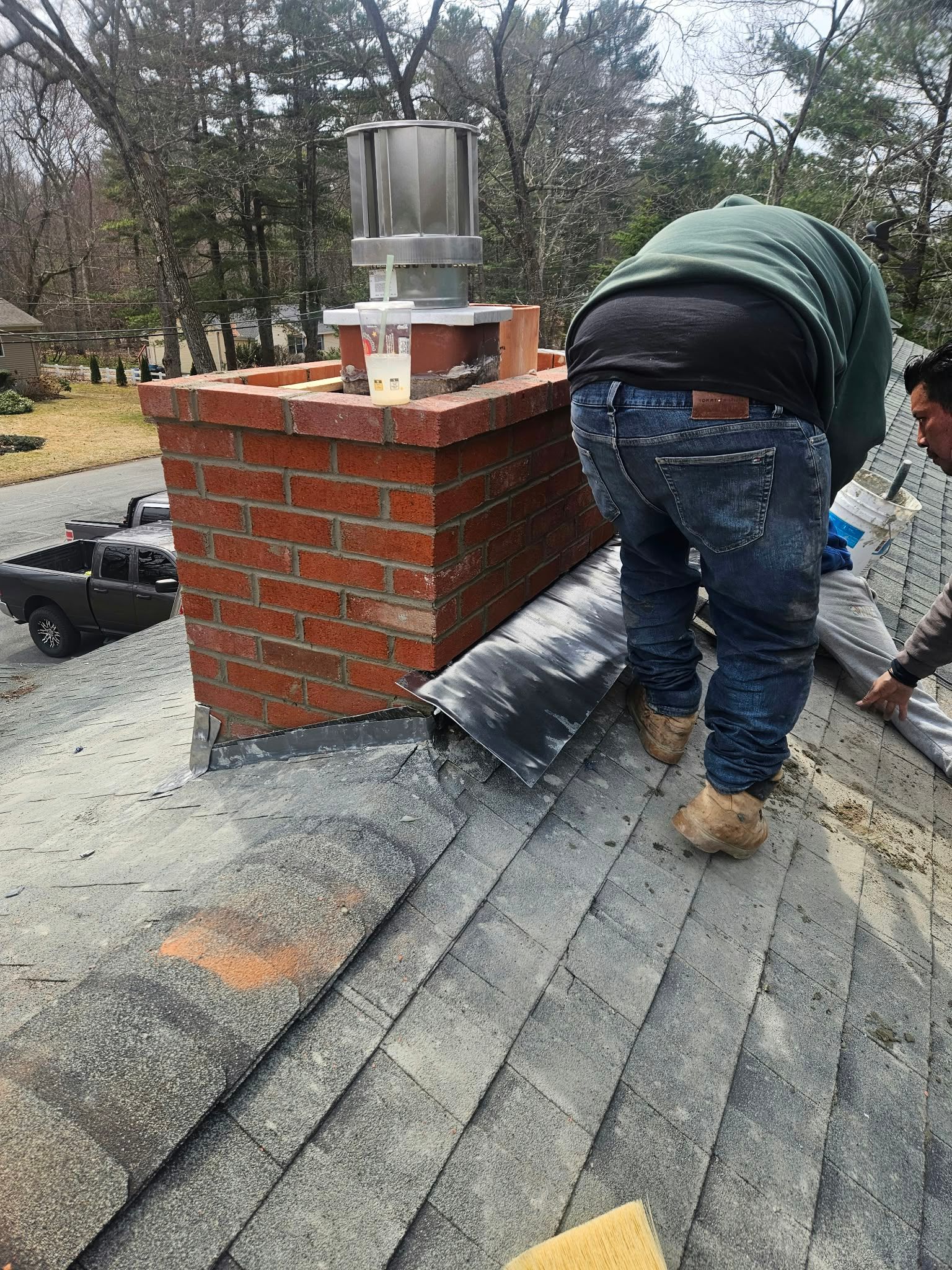 Worker repairing a shingled roof beside a brick chimney with metal cap and roofing materials.