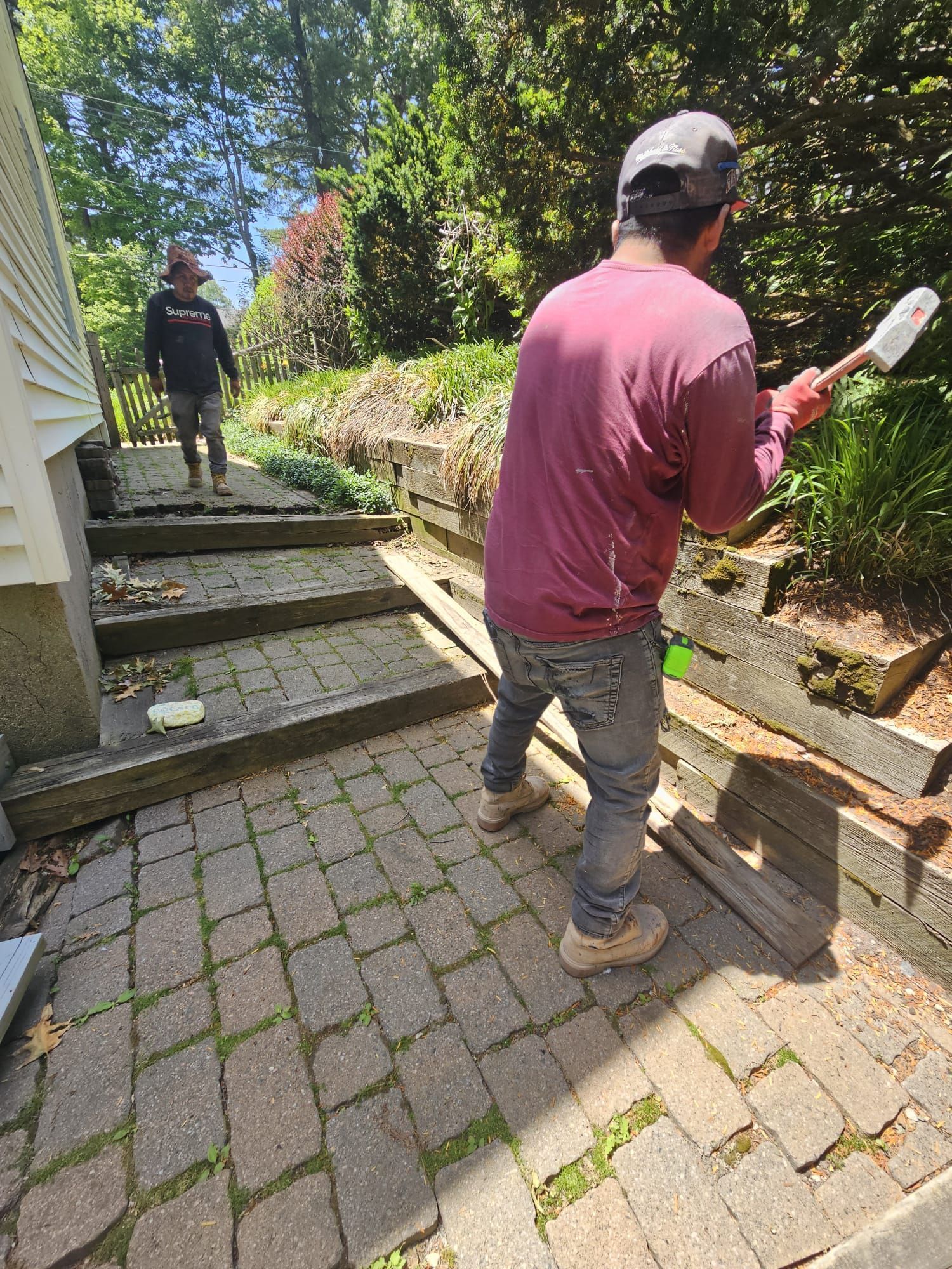 Two people using a brush and trowel to clean a brick walkway beside a house and garden bed
