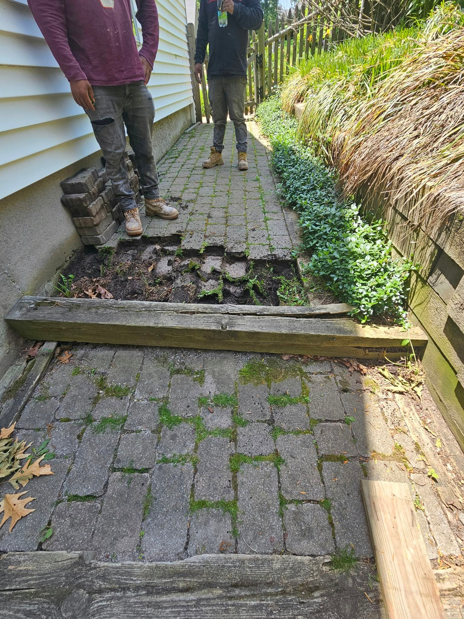 Two people standing on a narrow, broken stone path beside a house and garden slope