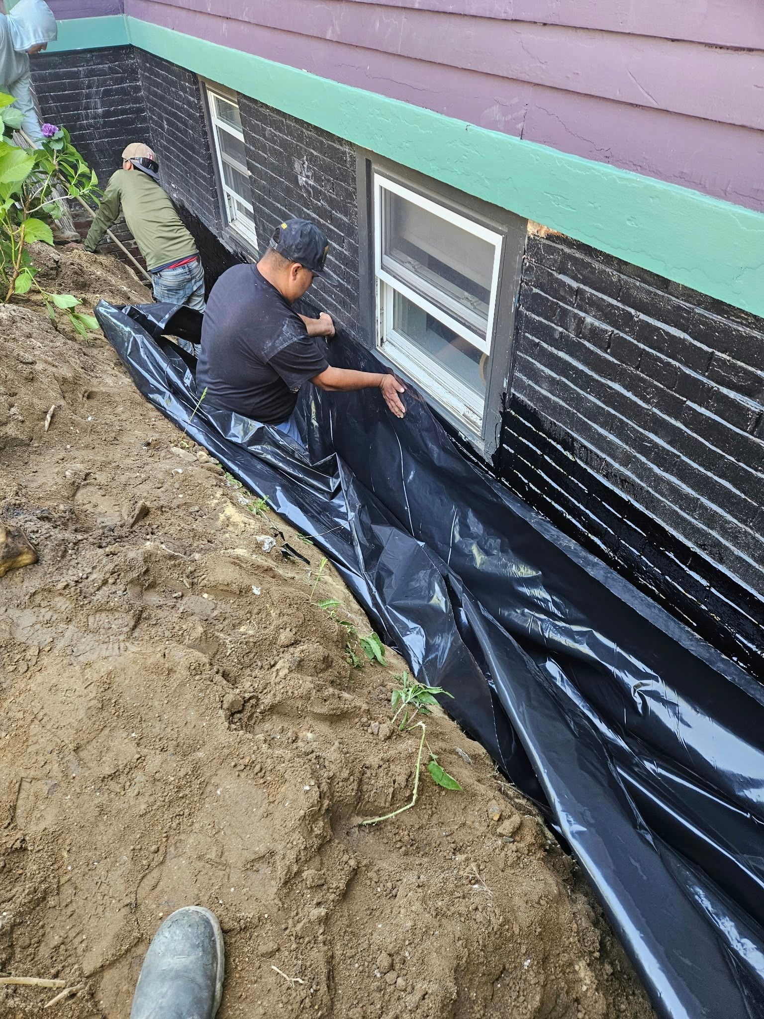 Workers installing black plastic sheeting along a house foundation beside a window