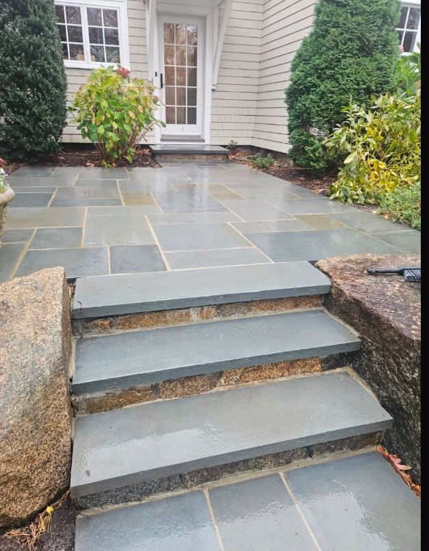 Stone patio steps leading to a white front door, bordered by shrubs and landscaping.