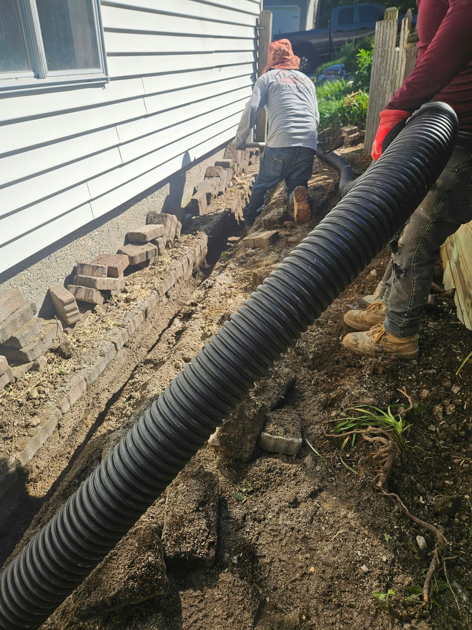 Workers laying black drainage pipe beside a white house; one child watches nearby.
