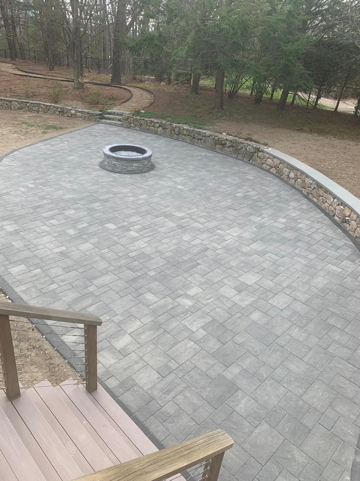 Gray paver patio with a round fire pit, bordered by trees and a wooden deck railing in the foreground