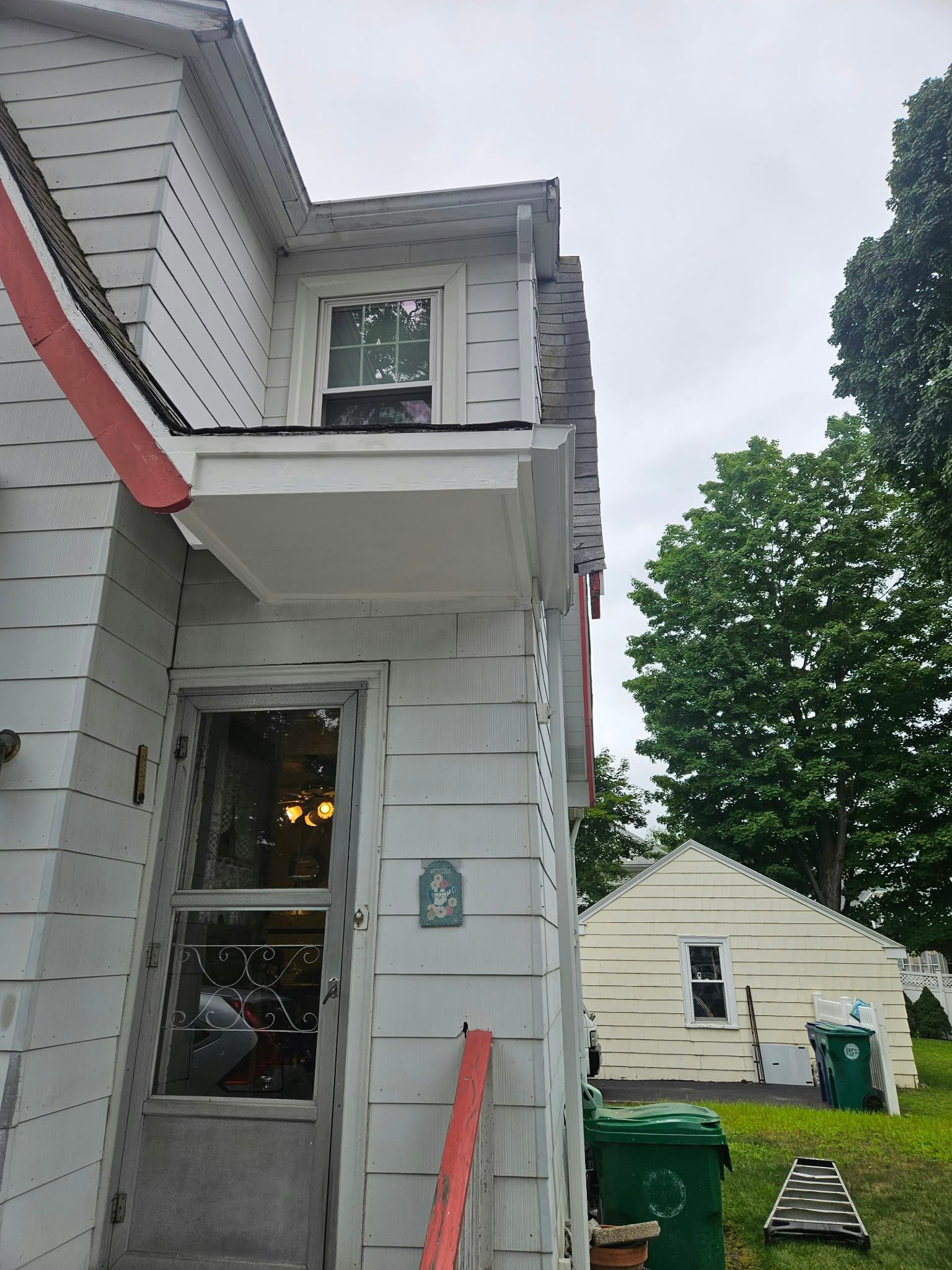Side of a gray house with a small porch, red trim, and a window above, next to a driveway and green bins.