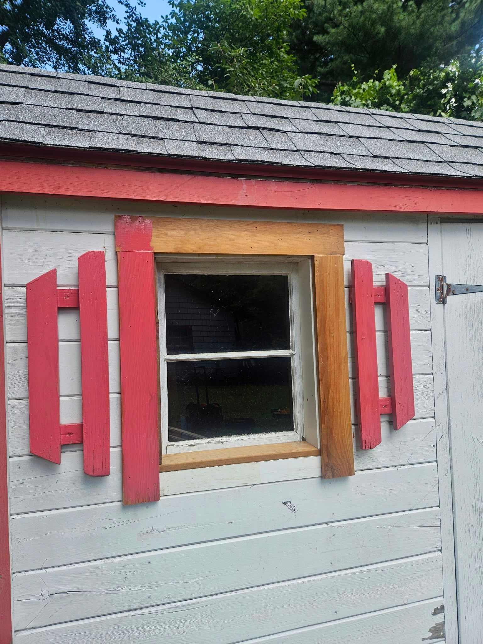 Small white shed wall with a central window framed by bright red shutters and tan trim.