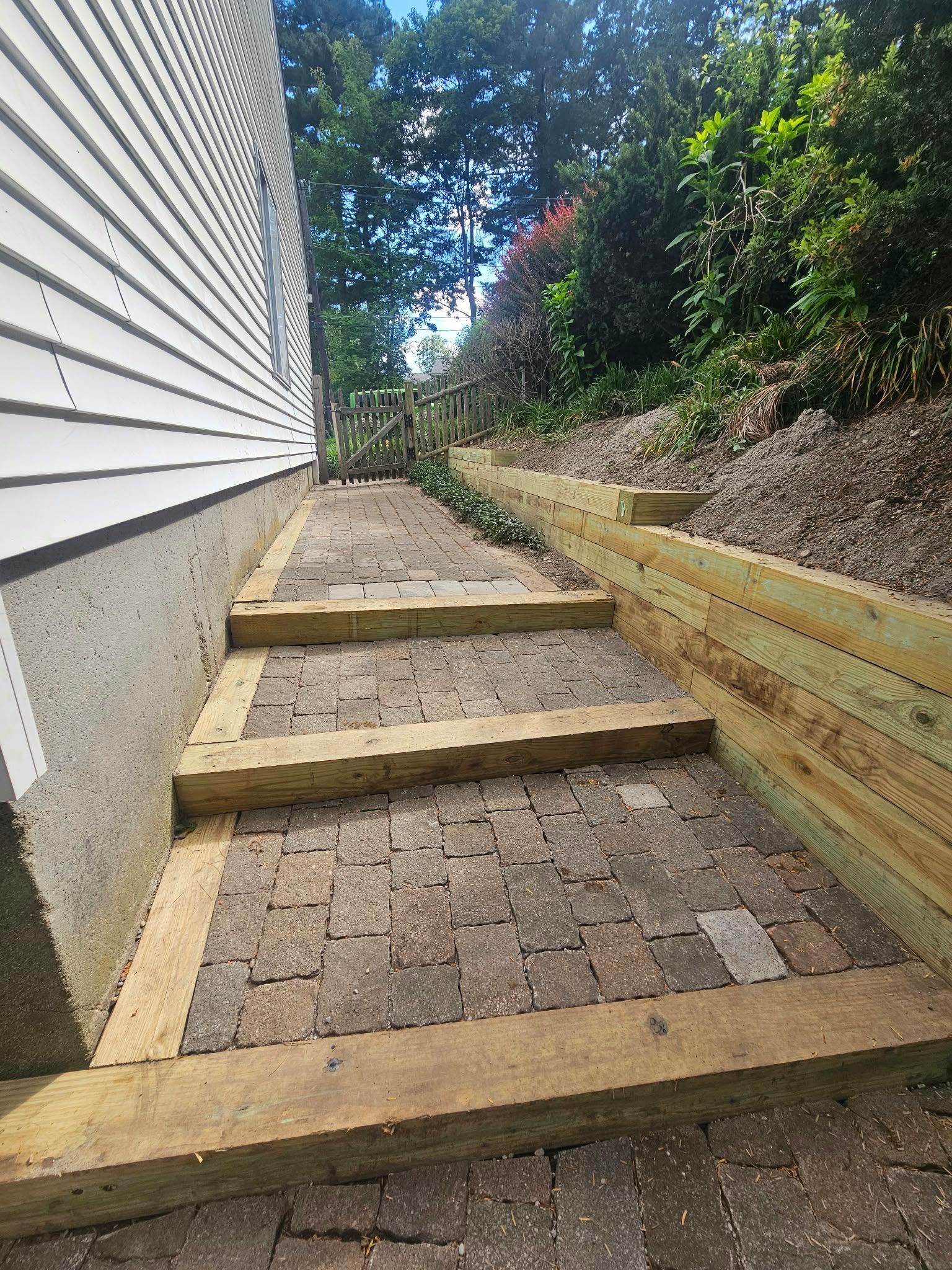 Narrow brick stairway beside a house, with wooden retaining walls and greenery on a slope.