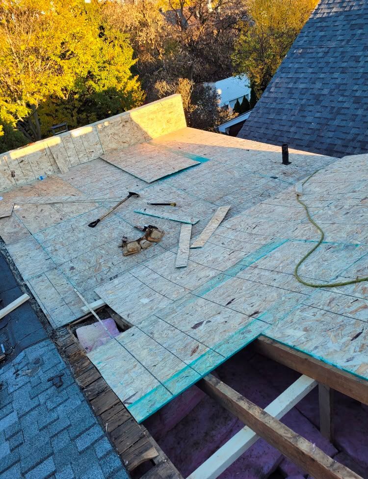 Rooftop under construction with plywood decking, blue chalk lines, and scattered tools beside shingles and trees