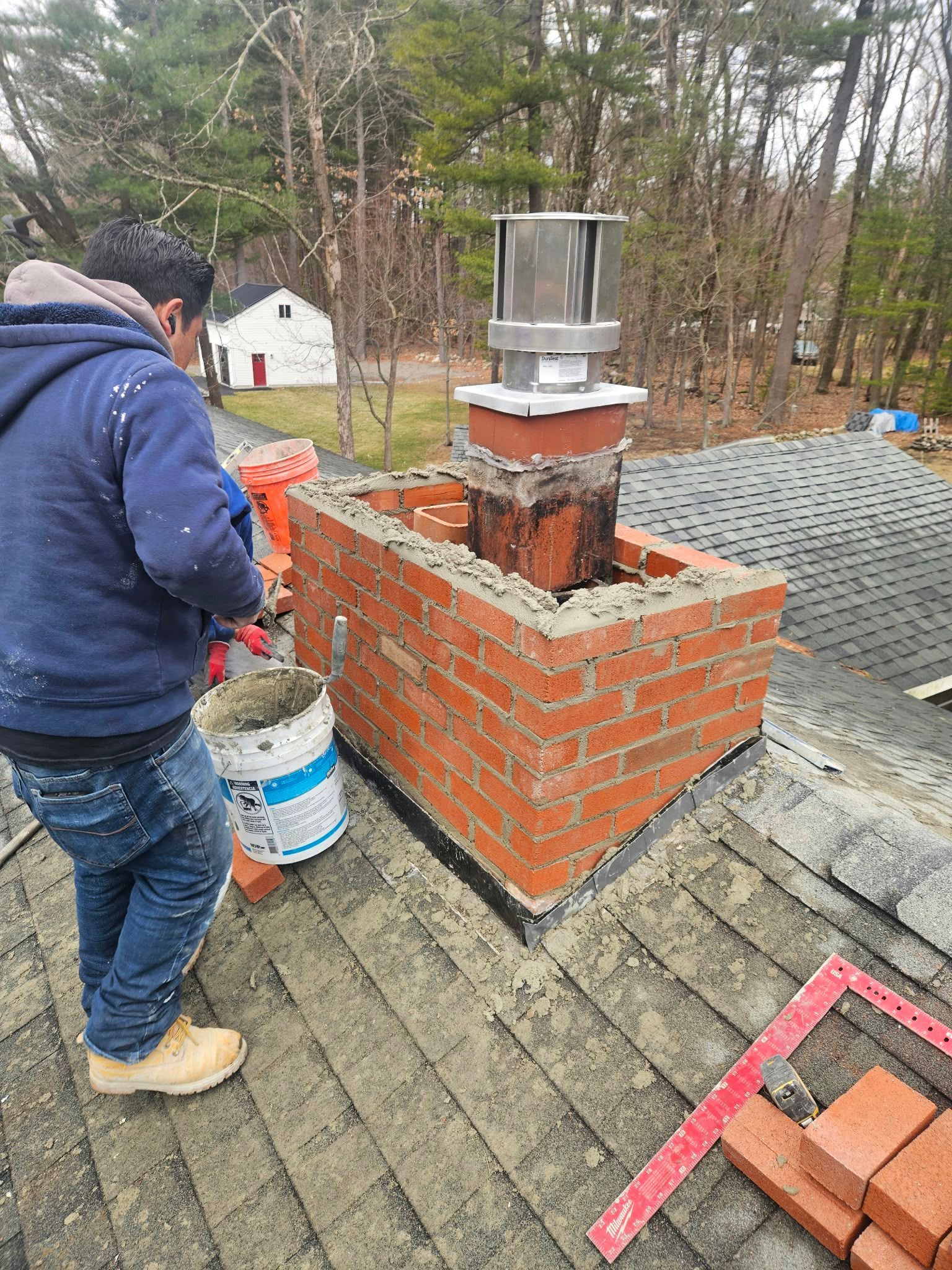 Worker troweling red brick chimney on roof with bucket and stacked bricks nearby