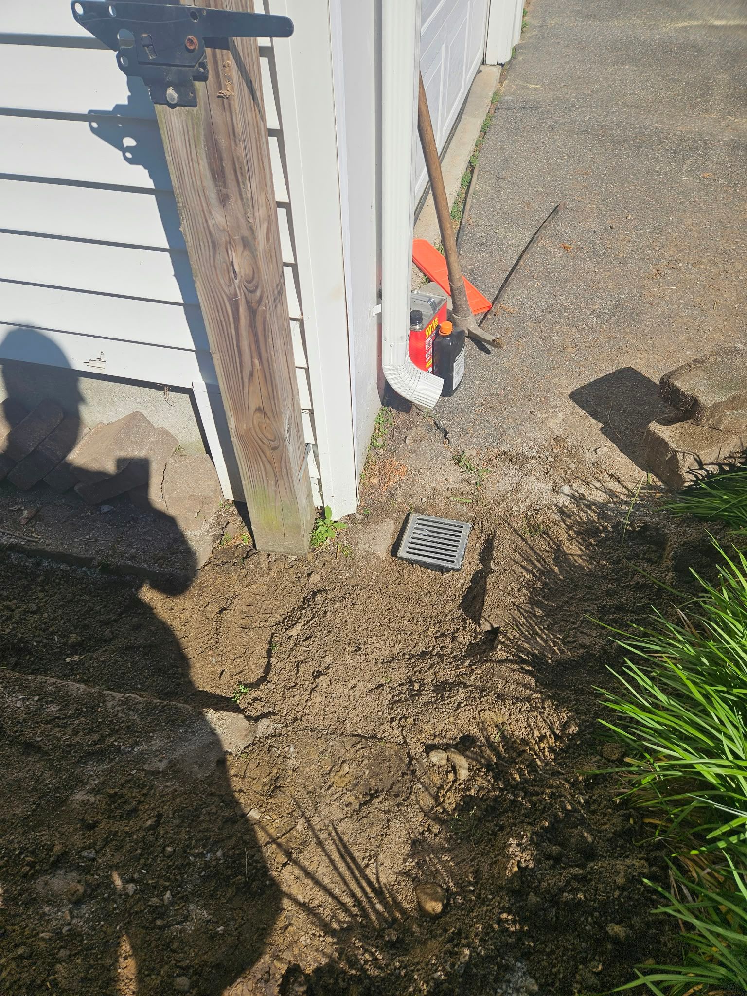 Corner of house exterior with dirt, a drain grate, and yard tools leaning against the wall.