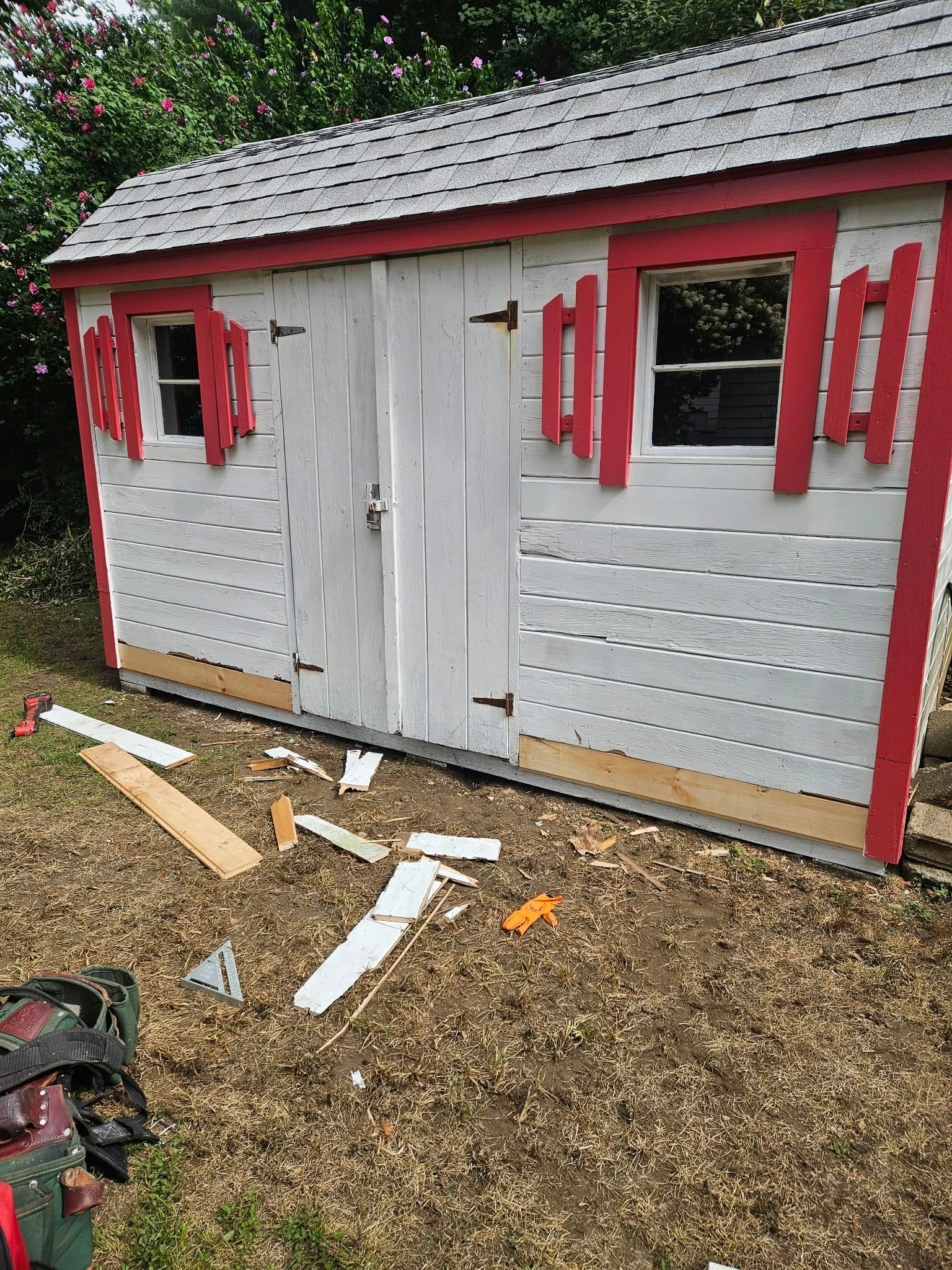 Small gray shed with red trim and a white door, sitting on wood chips with debris in front.