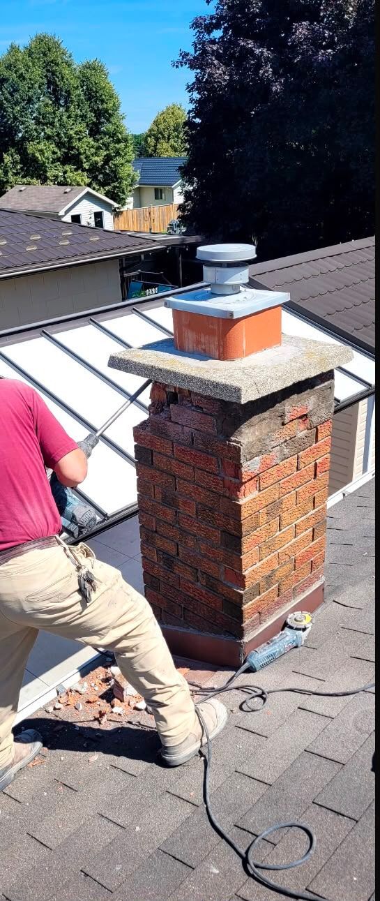 Worker on a shingled roof inspecting a brick chimney with a metal cap.