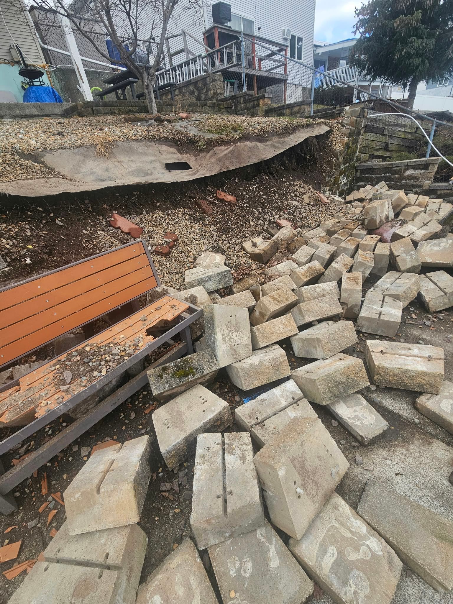 Collapsed retaining wall with scattered concrete blocks beside a house and metal steps.