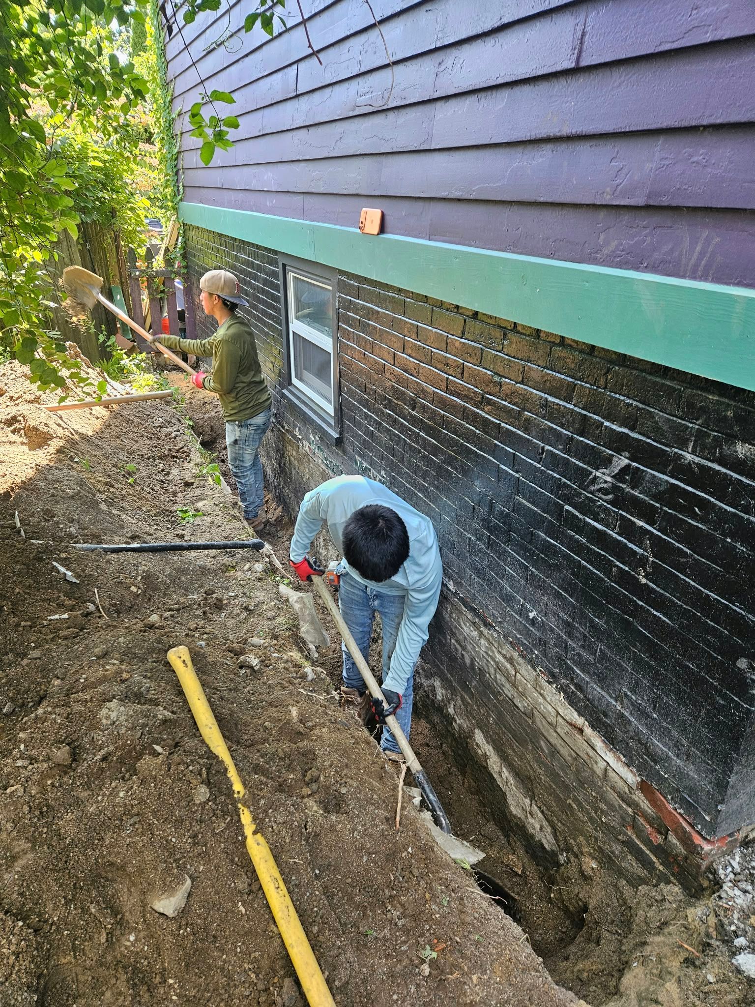Workers digging a trench beside a house with black and green siding on a wooded lot