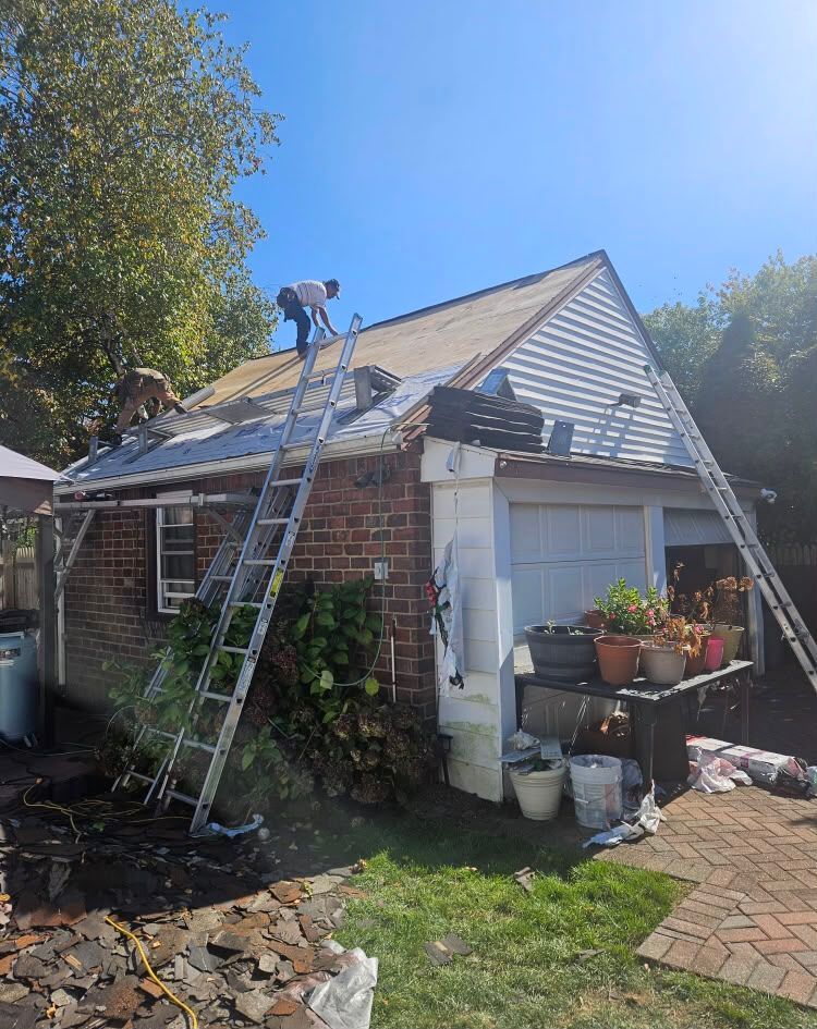 Two workers on ladders repairing a roof on a brick house beside a garden in sunny weather