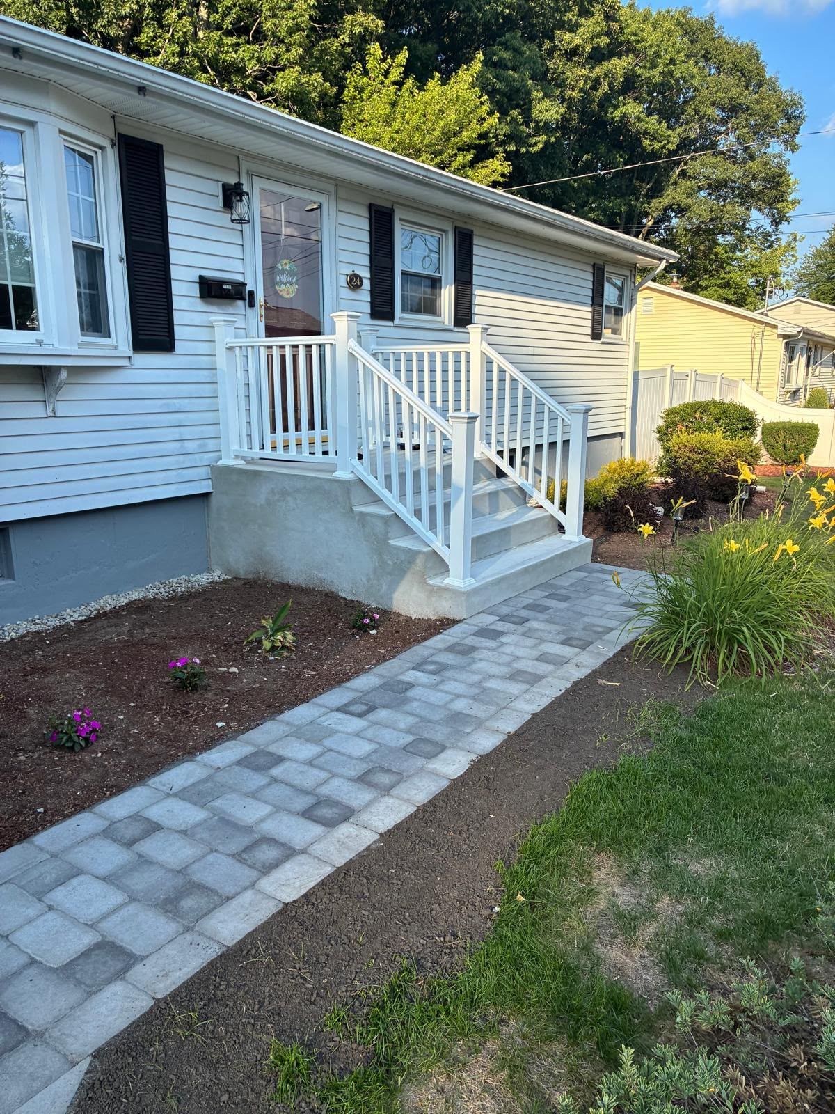 Front porch of a white house with steps, railing, and a paver walkway leading through a landscaped yard