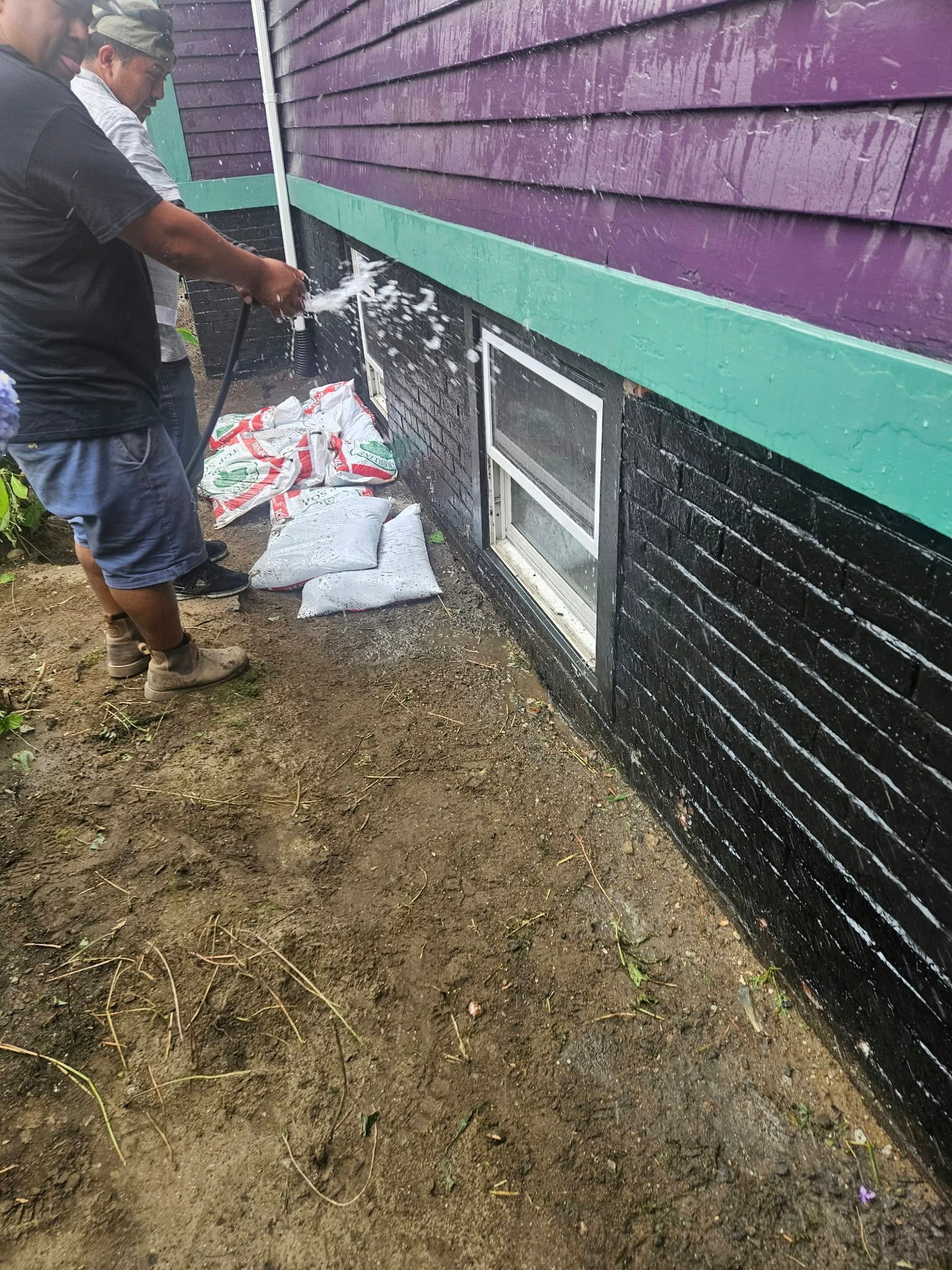 Worker beside house foundation spraying water along purple and black exterior wall in muddy yard