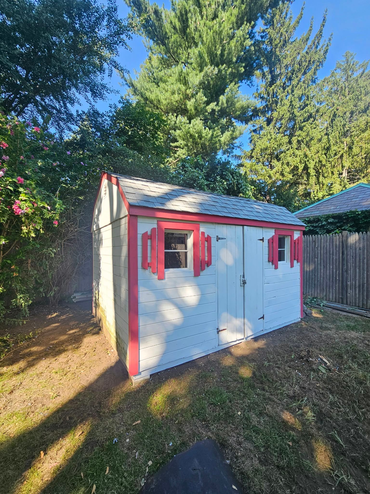 White garden shed with red trim and shutters in a sunny backyard, surrounded by trees and grass.