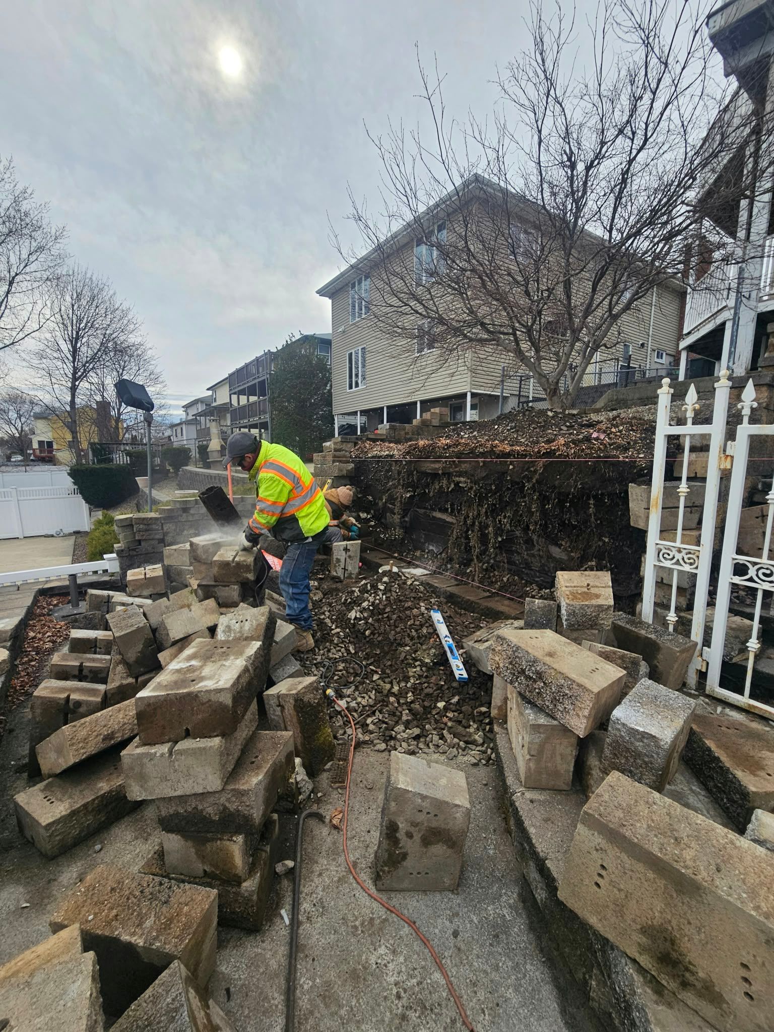 Worker in neon jacket clearing broken stone blocks from a muddy residential walkway.
