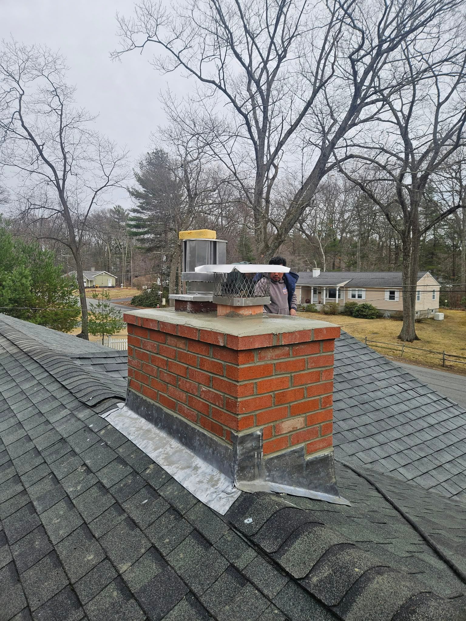 Brick chimney on a shingled roof with a person working beside it on a cloudy day