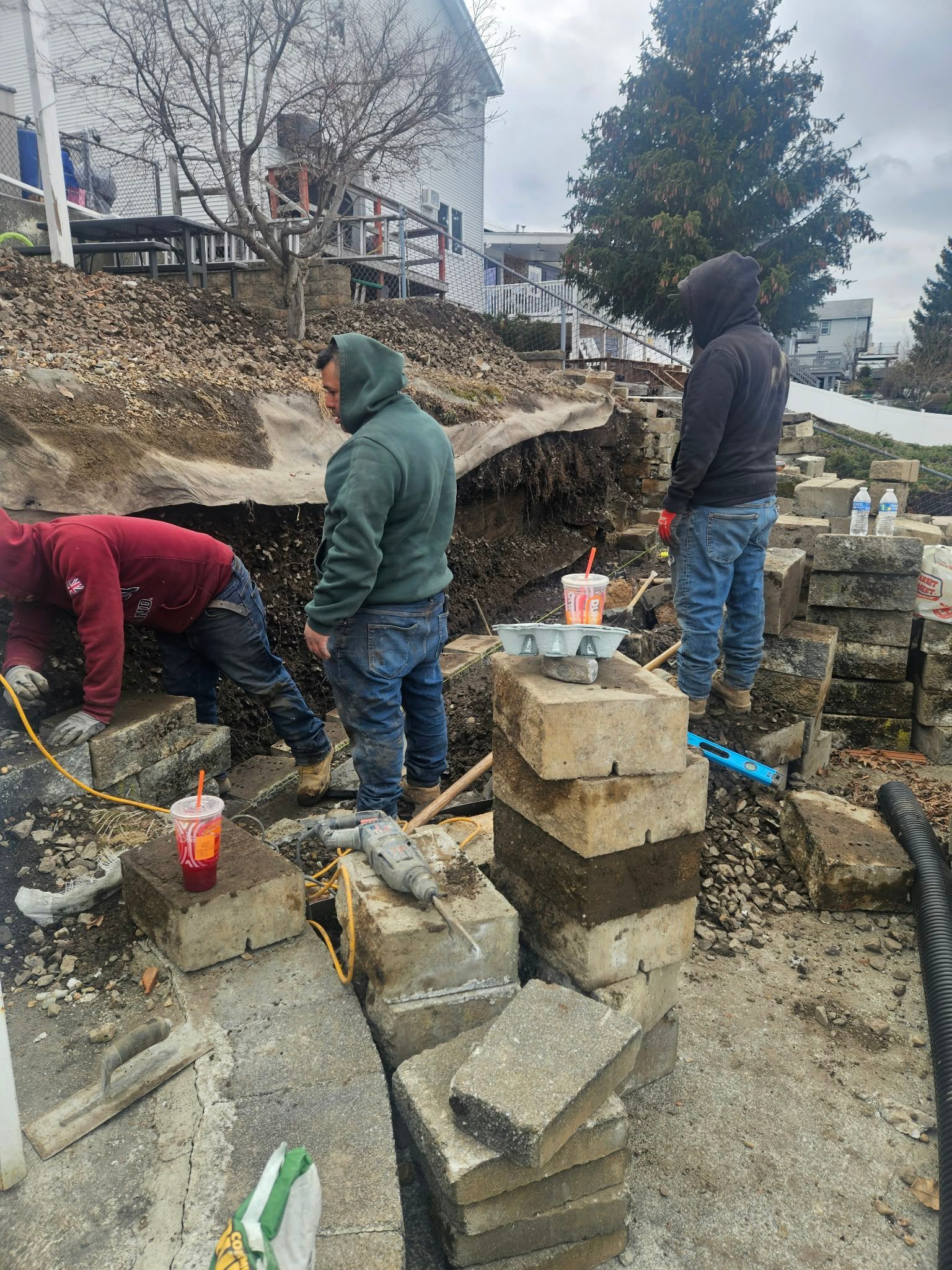 Workers in hoodies digging at a construction site beside stacked concrete blocks and a tree