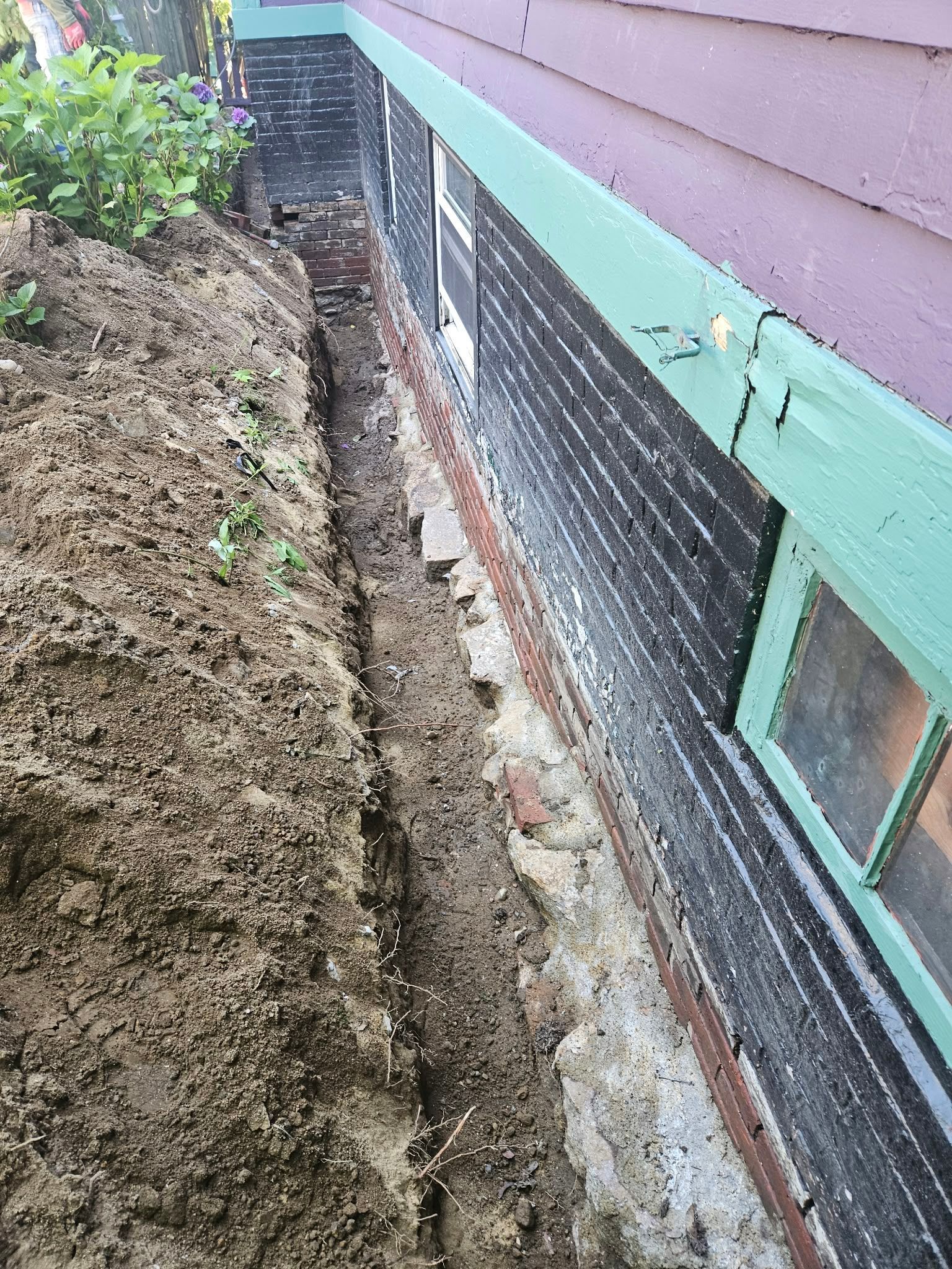 Narrow dirt trench beside a house foundation with exposed black waterproofing and green siding.