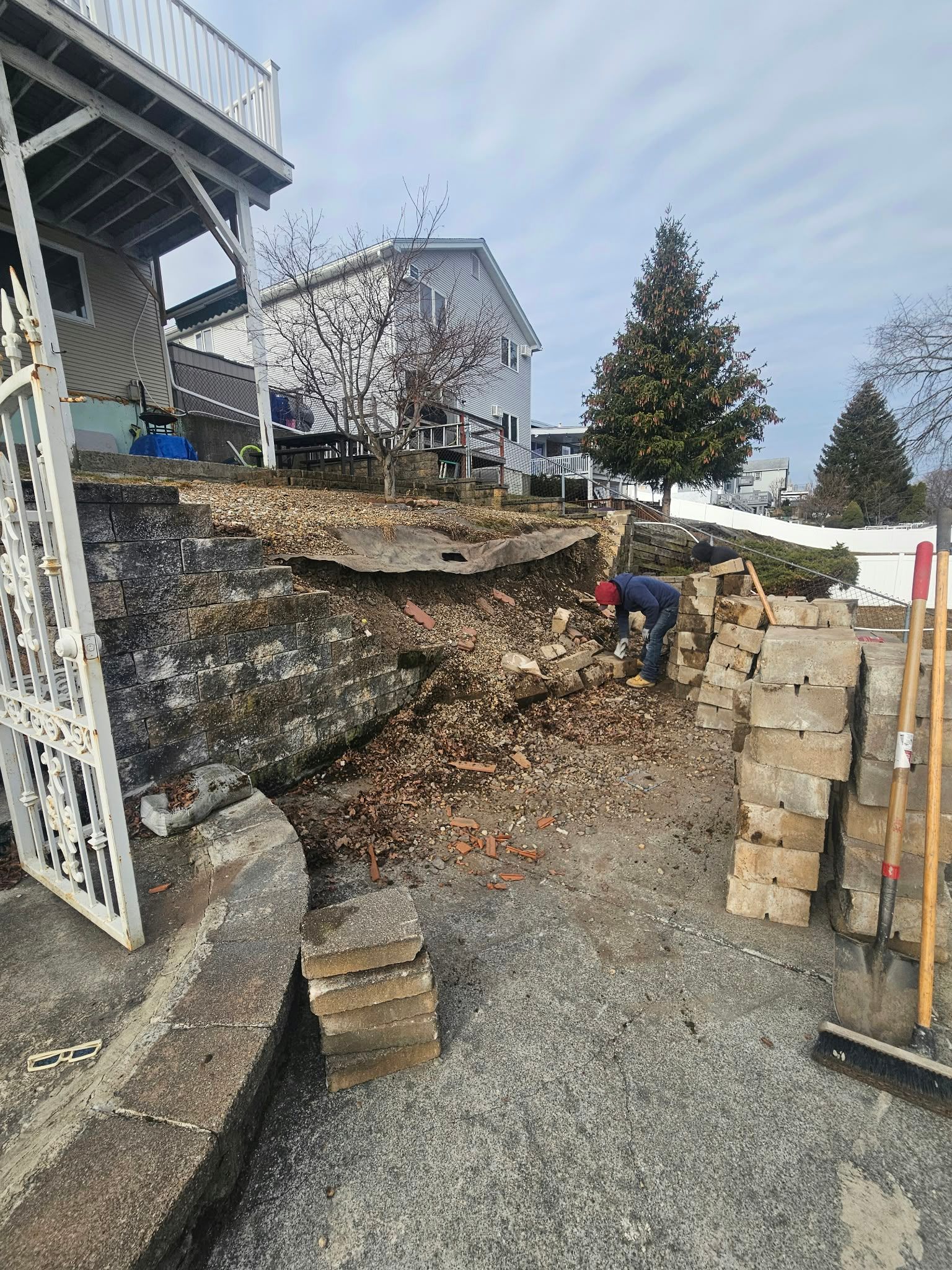 Landslide cleanup beside a house, with workers near a steep soil embankment and stacked retaining blocks.