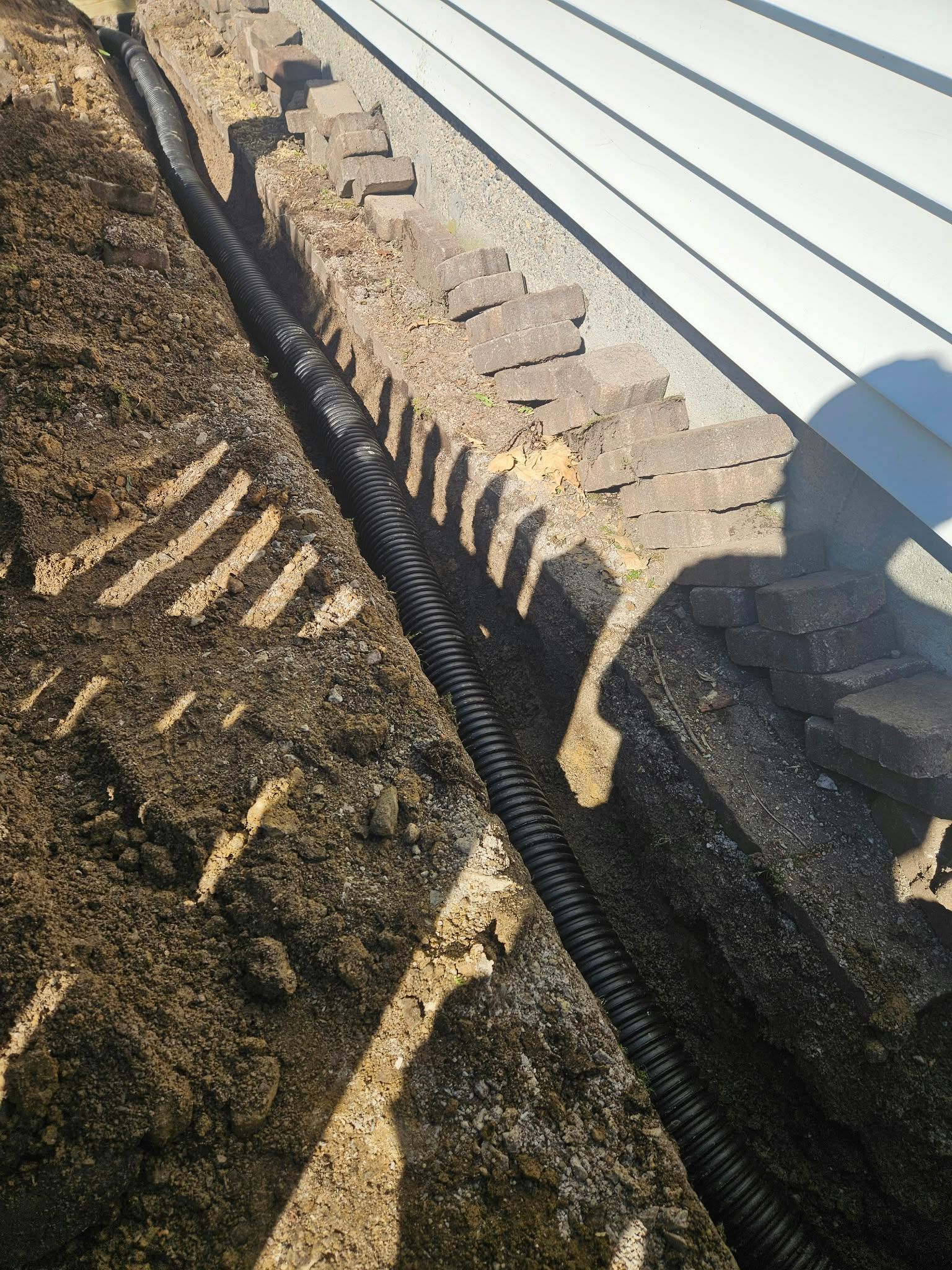 Landscape fabric-lined trench beside white siding with stacked stones and gravel edge