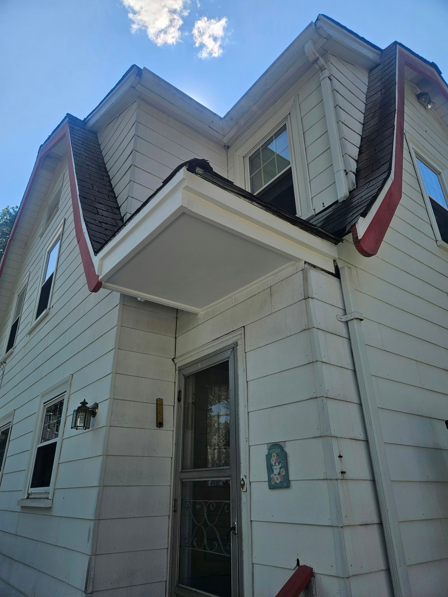 White two-story house with steep roof, red trim, and a small front porch against a blue sky