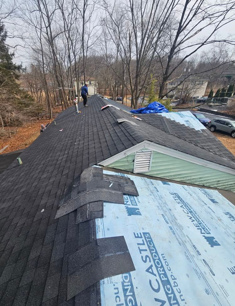 Roofers repairing a shingled roof with underlayment exposed on a wooded residential home