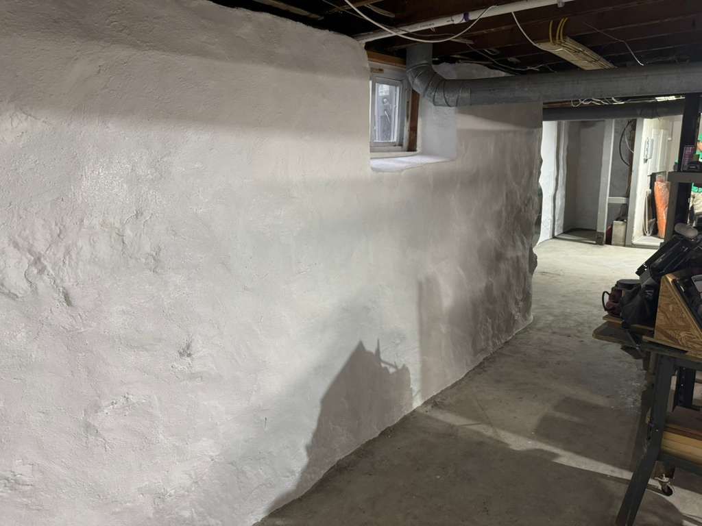Narrow basement hallway with white plaster walls, exposed ceiling beams, and a doorway at the far end.