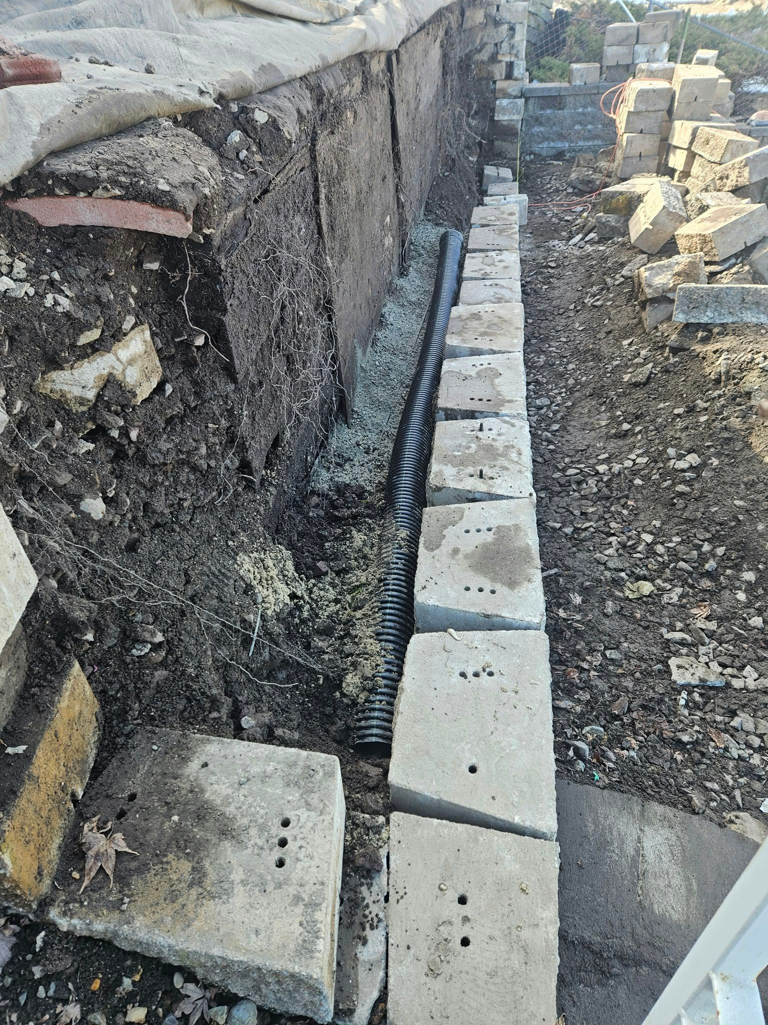 Narrow trench with gravel, concrete blocks, and black drainage mesh along a stone wall under construction