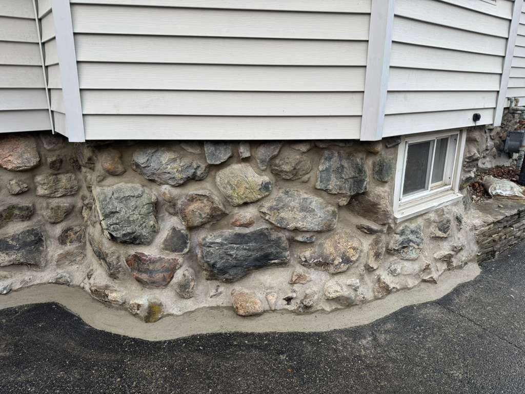 Stone foundation with small basement window beneath white siding beside an asphalt driveway
