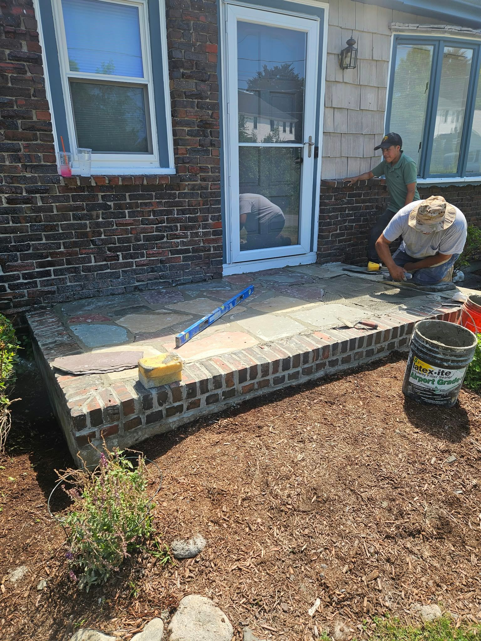 Two workers repairing a front porch and steps beside a house, with tools and a bucket on the ground