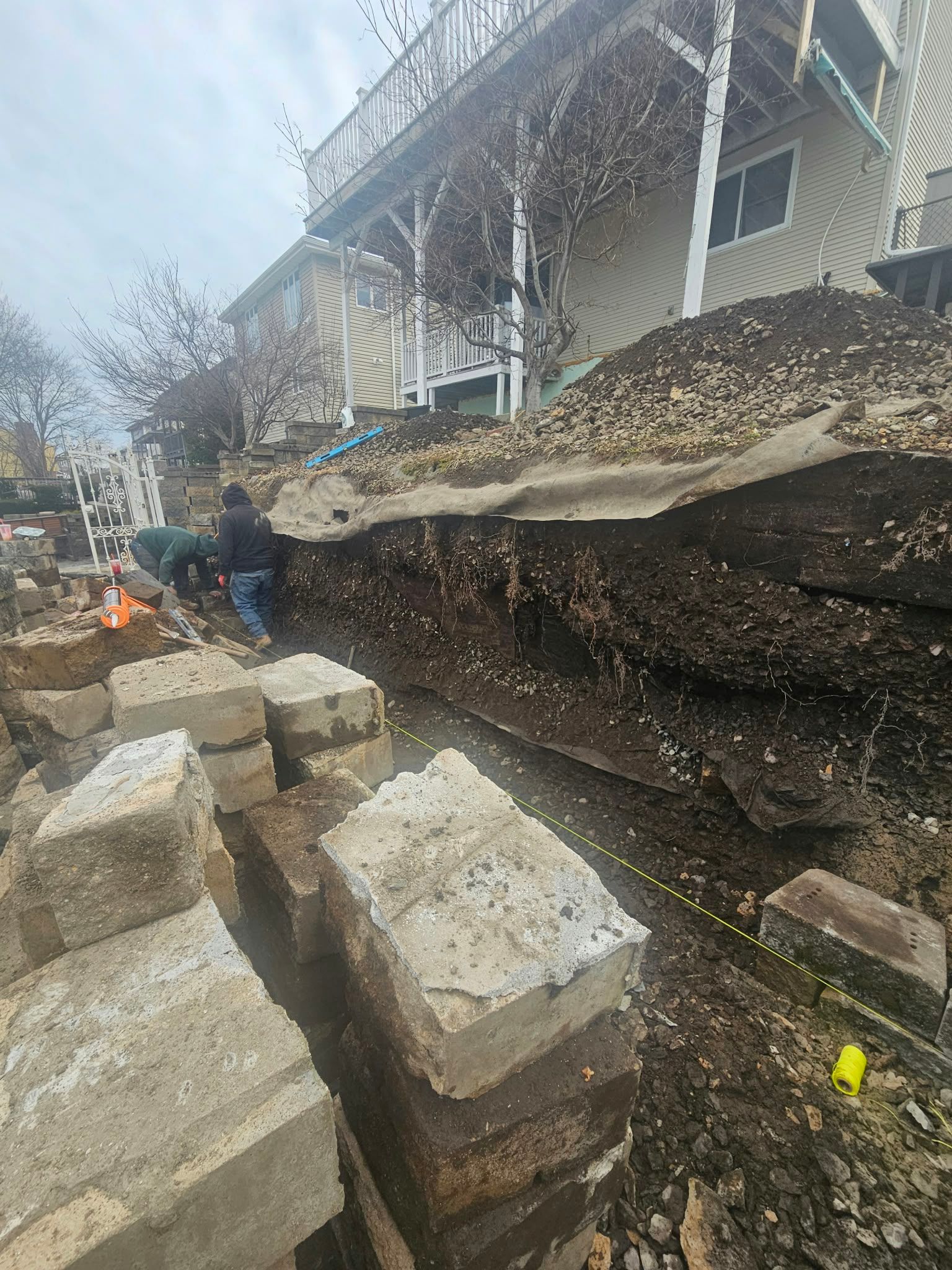 Collapsed sidewalk beside a house, with large broken concrete slabs and a narrow trench along the foundation.