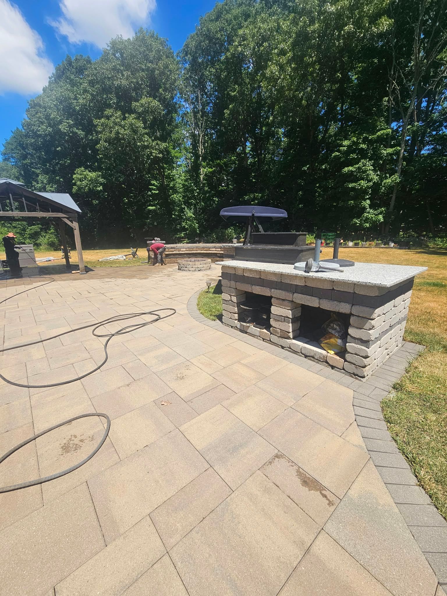 Outdoor patio with stone pavers, a built-in grill, and trees in the background on a sunny day