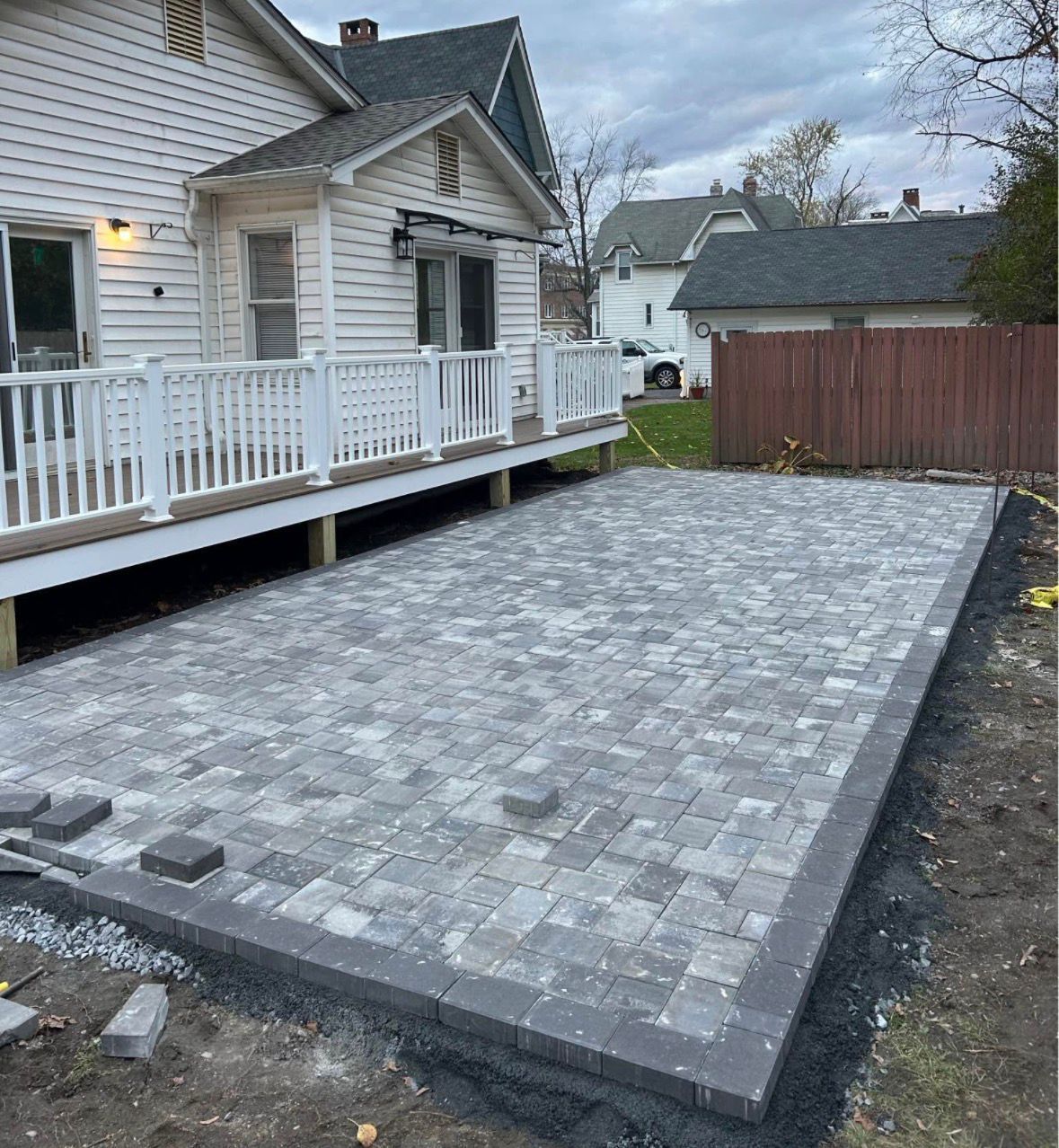 New gray paver patio beside a white house and deck, bordered by dark edging near a wooden fence