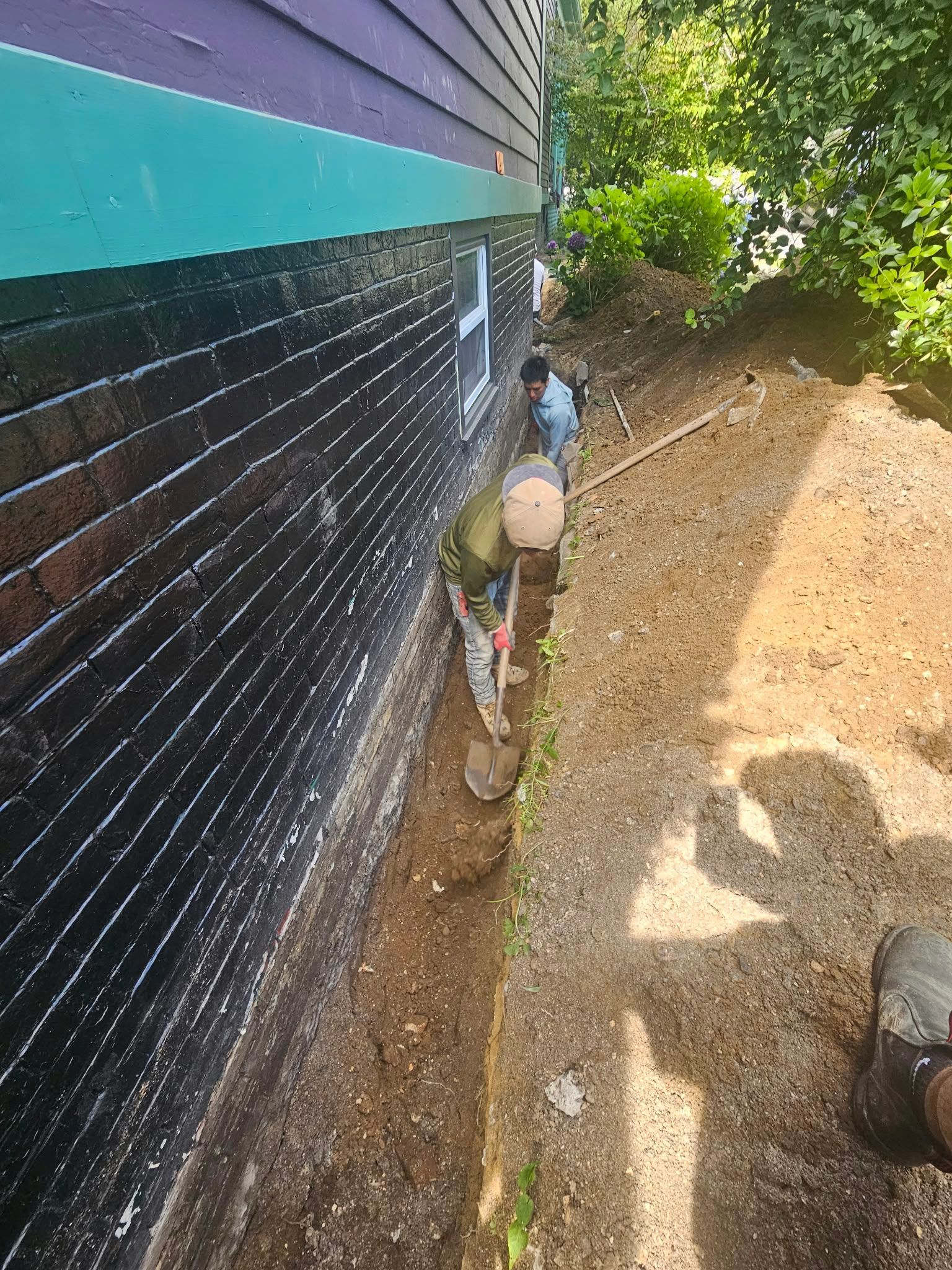 House siding with black and teal exterior, narrow gravel path, and garden hose beside a sunlit yard.