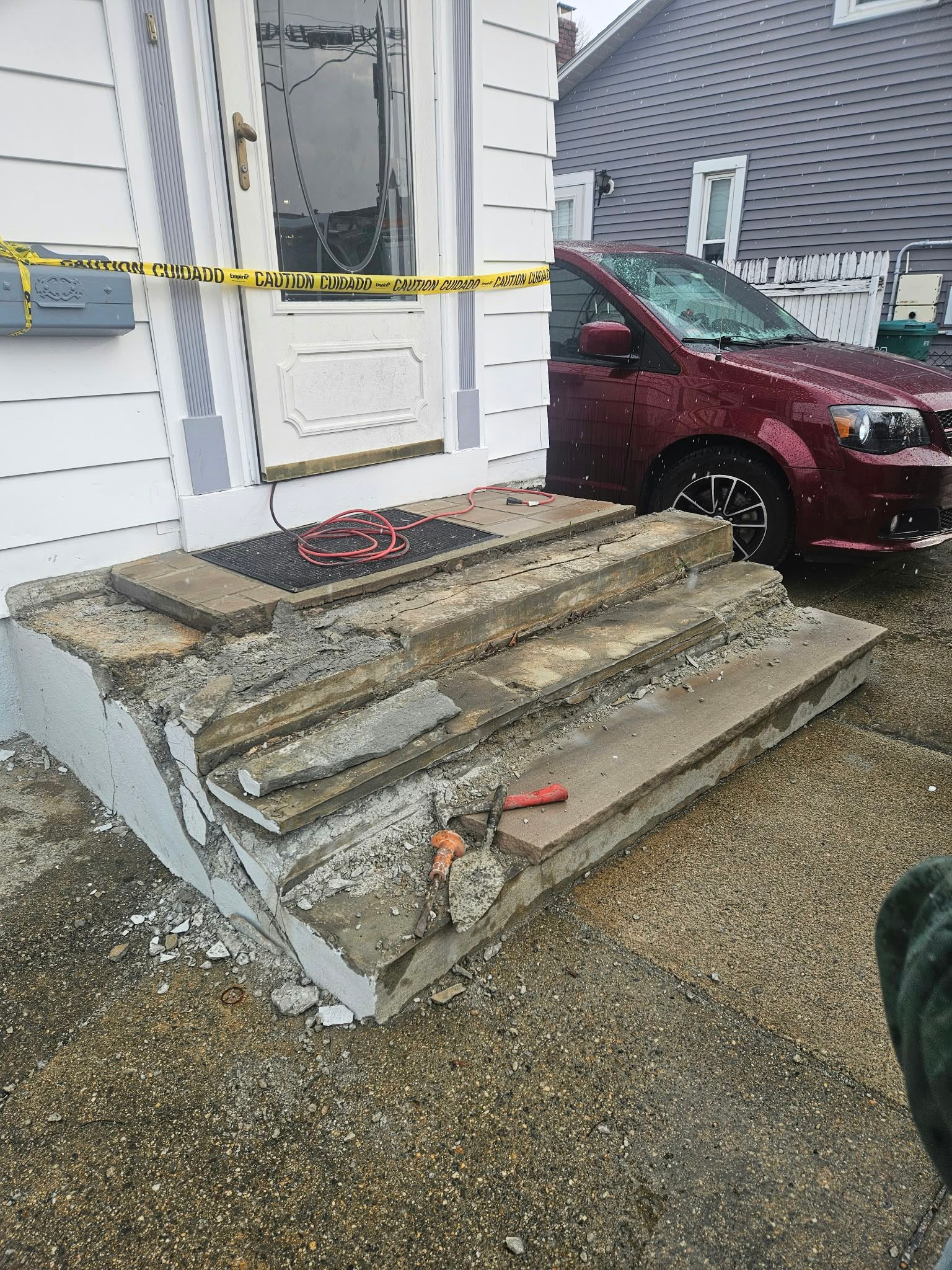 Damaged front steps taped off with caution tape beside a maroon car and white house doorway