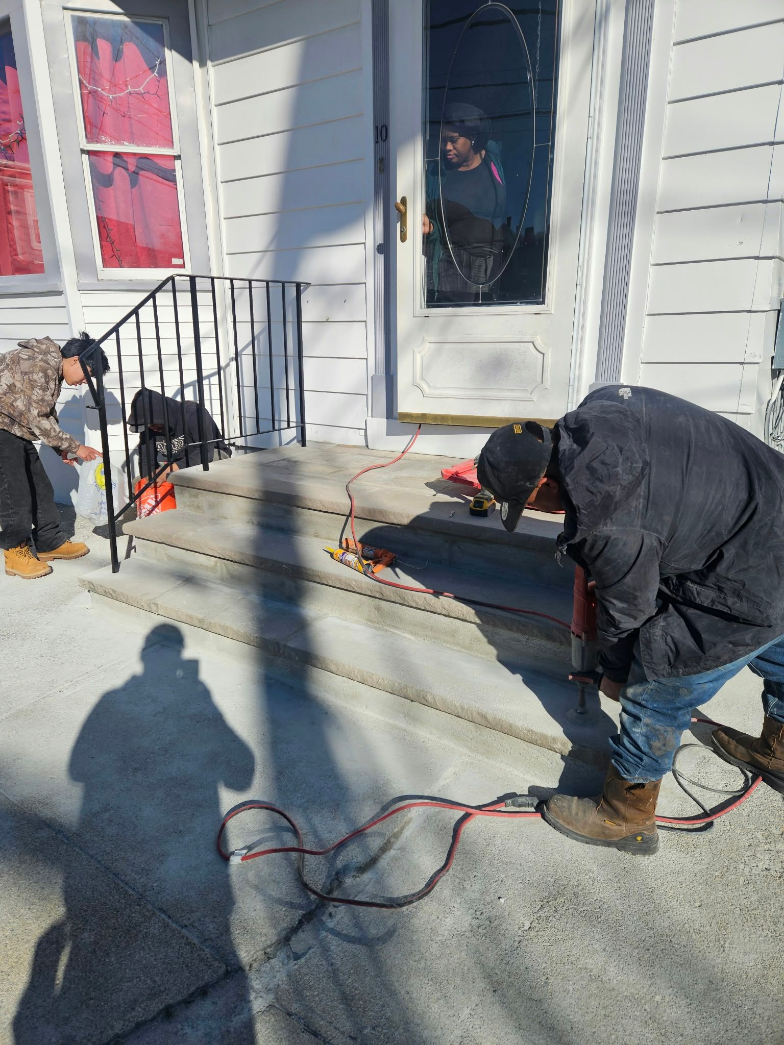 Two people kneel on a porch, sanding steps with power tools outside a white house.