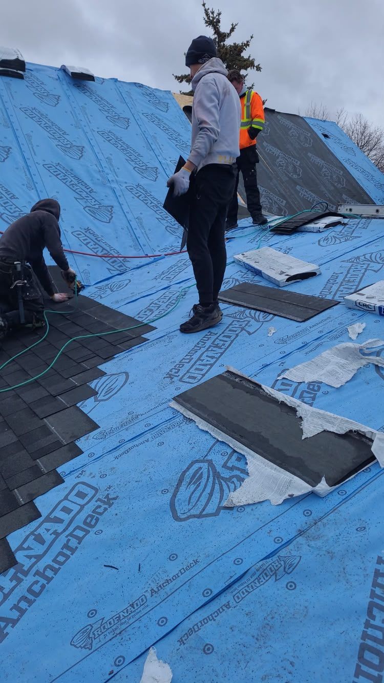 Workers installing roofing on a house, with blue underlayment and dark shingles in progress