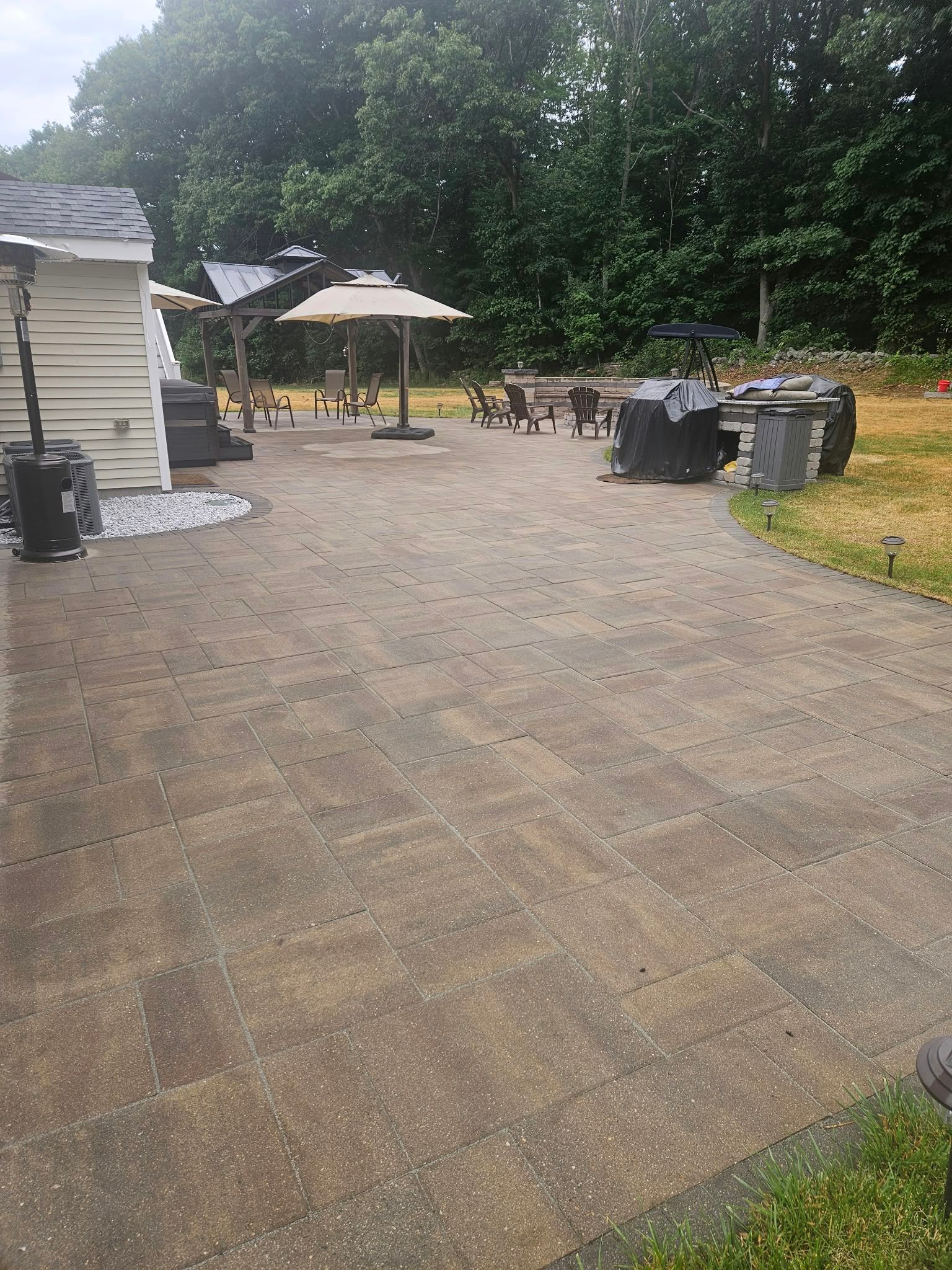 Outdoor patio with stone pavers, a gazebo, tables and chairs, and trash bins beside a white building and trees